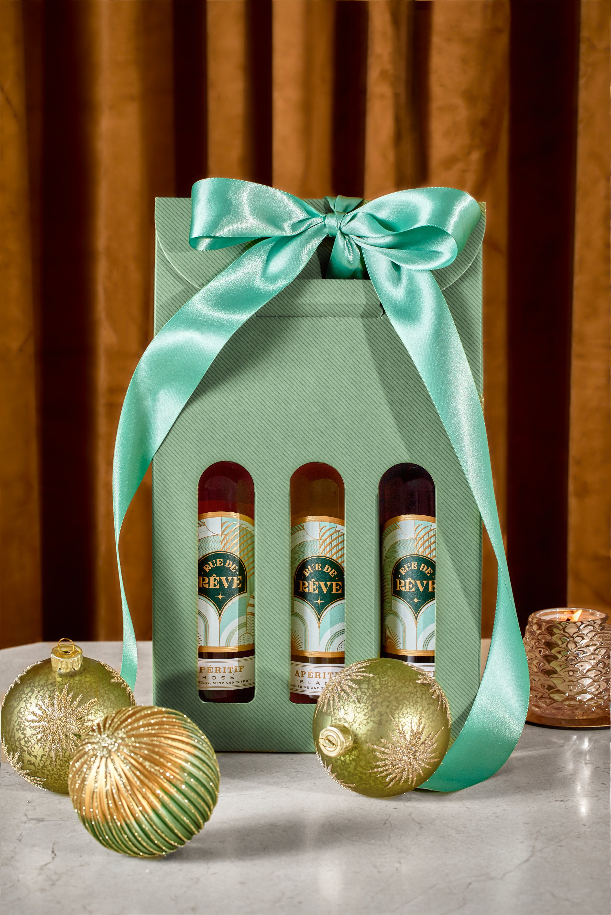 Gift set with three bottles of Rue de Rêve aperitif in a green box with a satin ribbon, surrounded by Christmas ornaments and a candle holder on a table with a brown curtain background.