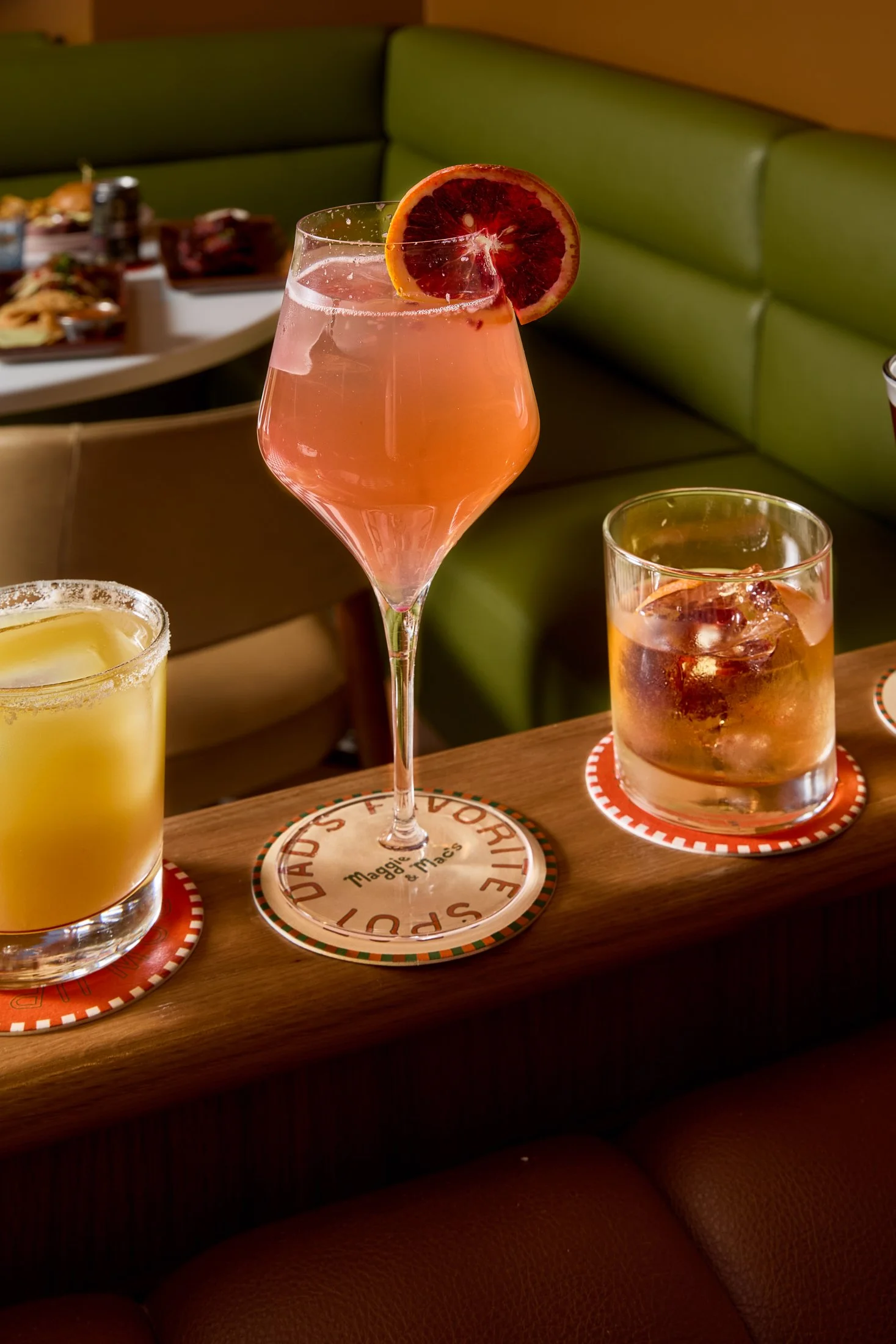 A pink cocktail with a slice of blood orange garnish on a dinner table, surrounded by other drinks, in a restaurant setting.