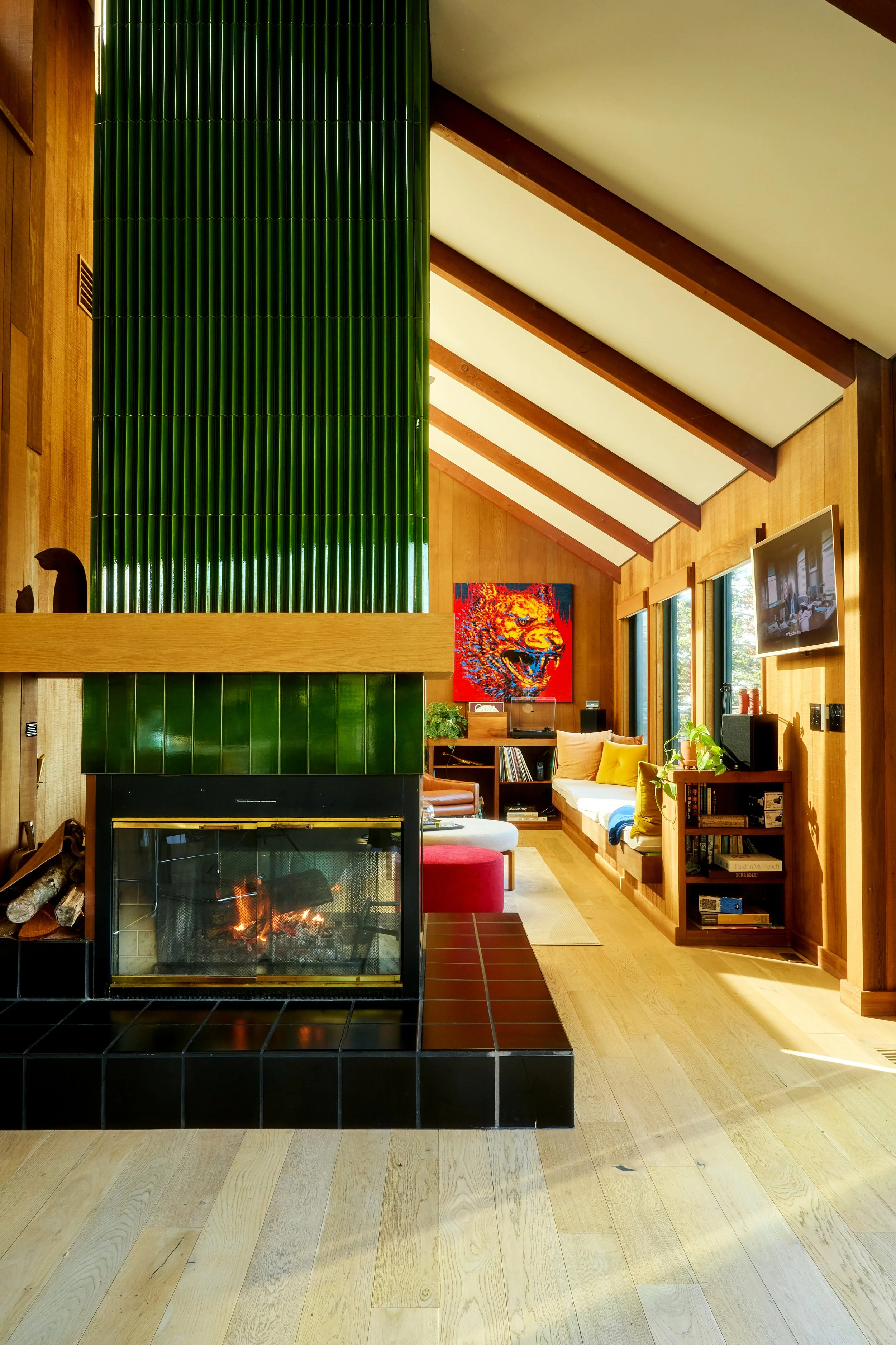 Living room with wood-paneled walls and ceiling, large green tiled fireplace, and a cozy seating area with a colorful painting on the wall.