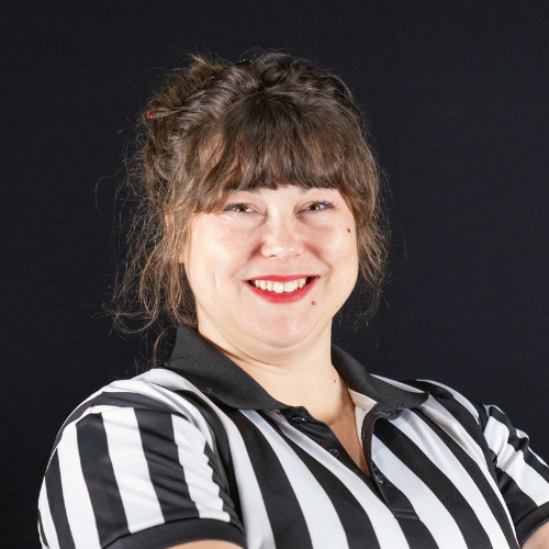 A woman with brown hair in a messy bun, wearing a black and white striped referee shirt, smiling at the camera against a black background.