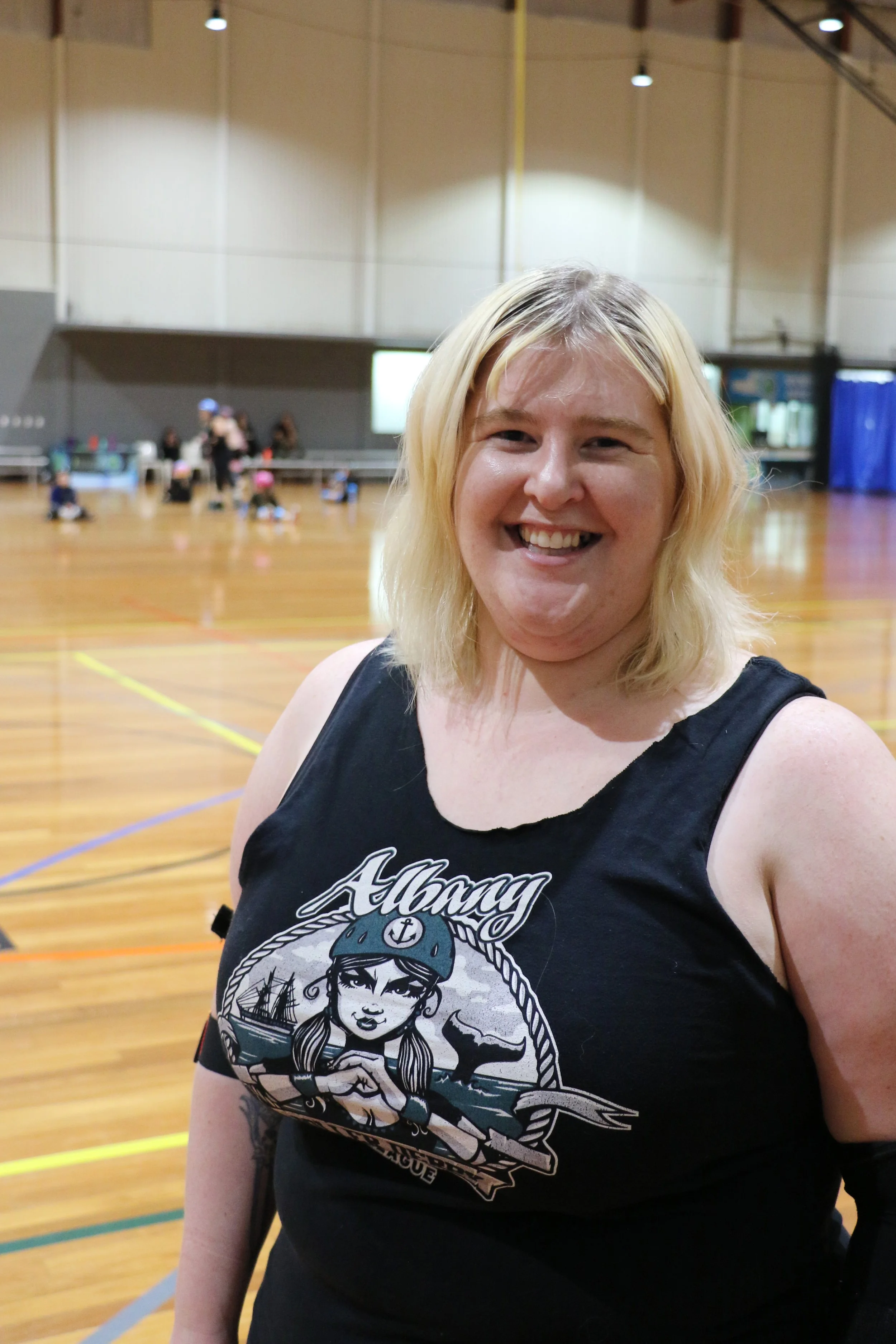 A smiling blonde person wearing a black tank top with a graphic that says 'Albany'  standing in an indoor sports facility with a wooden floor and blurred background of children and adults.