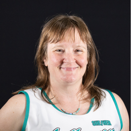 A woman with shoulder-length light brown hair, smiling, wearing a white athletic tank top with teal accents and text, against a black background.