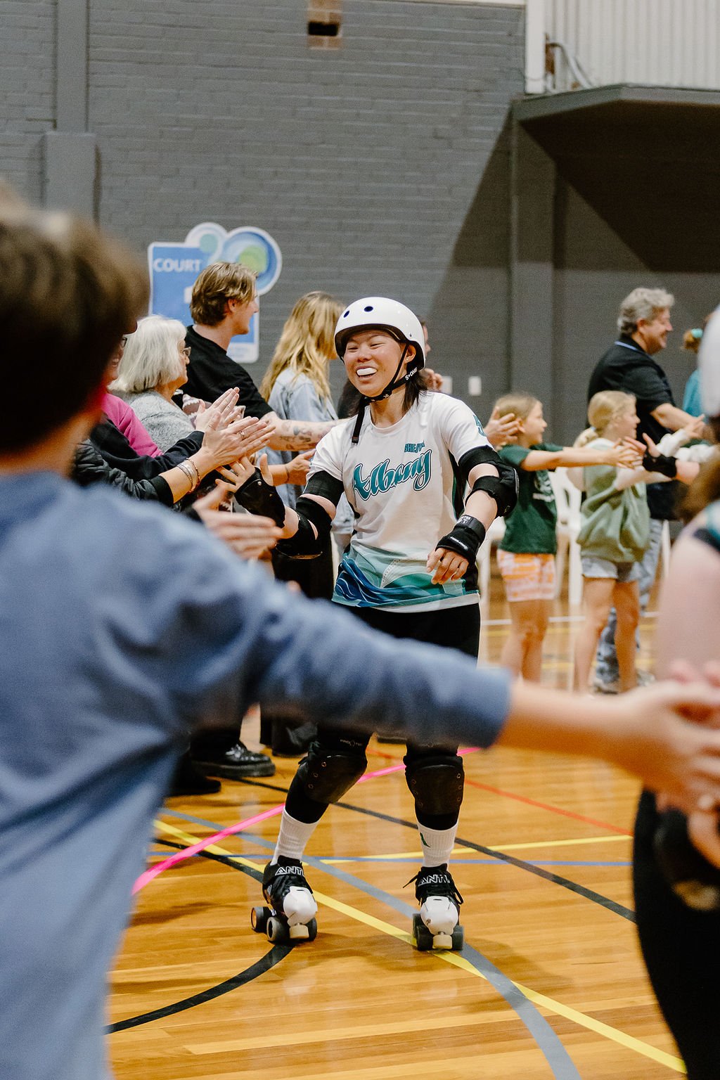 A woman on roller skates wearing a helmet and safety pads, smiling, receiving a high five from a crowd in a gymnasium with children and adults.