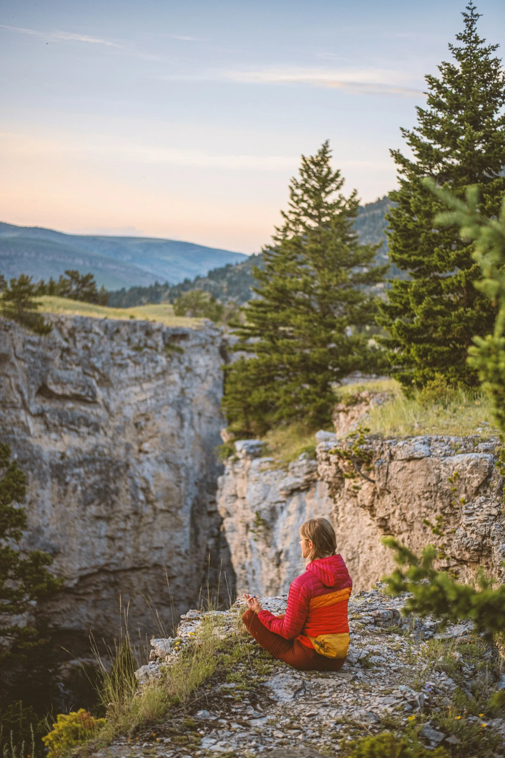 Woman in orange jacket sitting on edge of cliff admiring a canyon