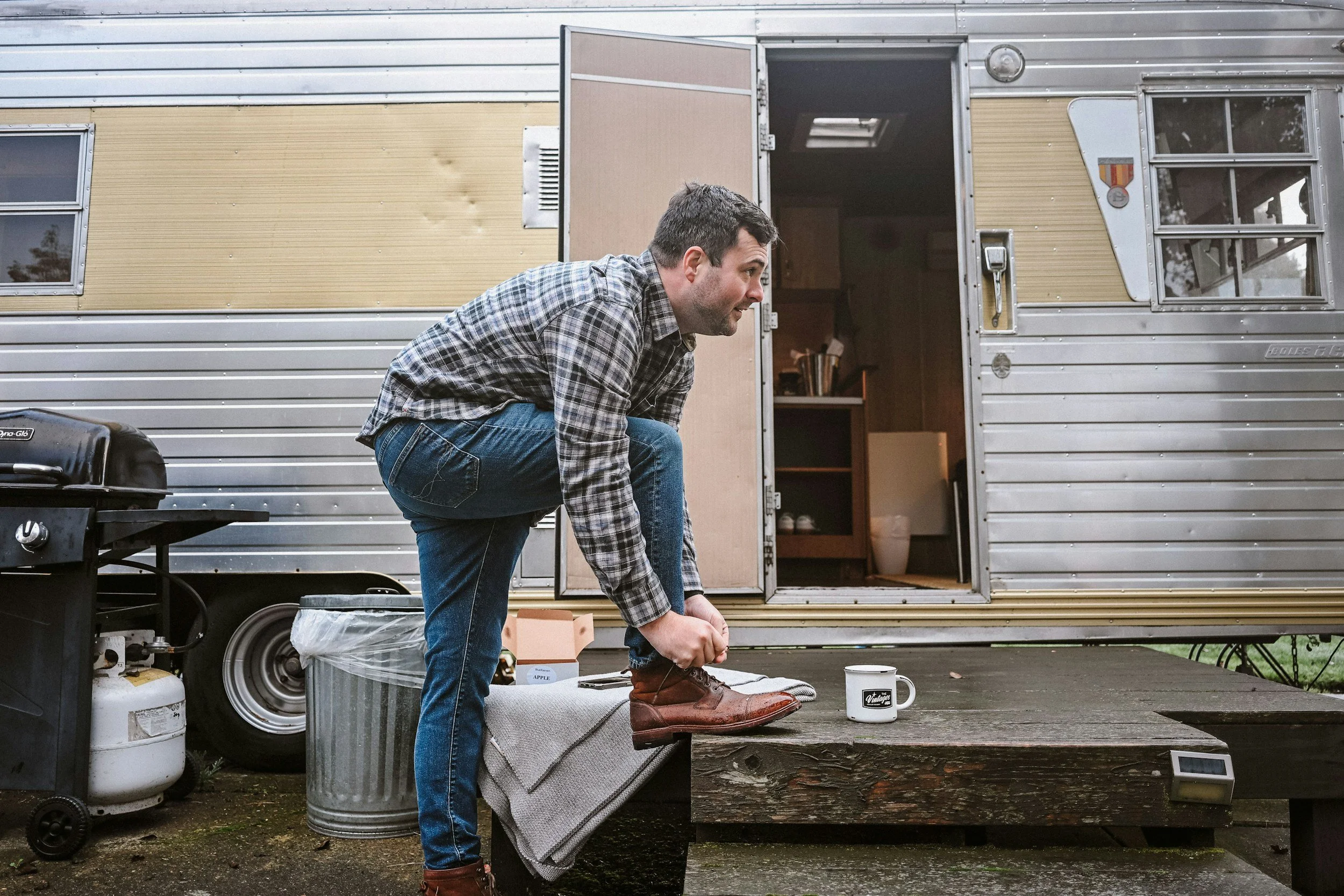 A man in a plaid shirt and jeans bending down to tie his shoe outside a silver and beige mobile home.