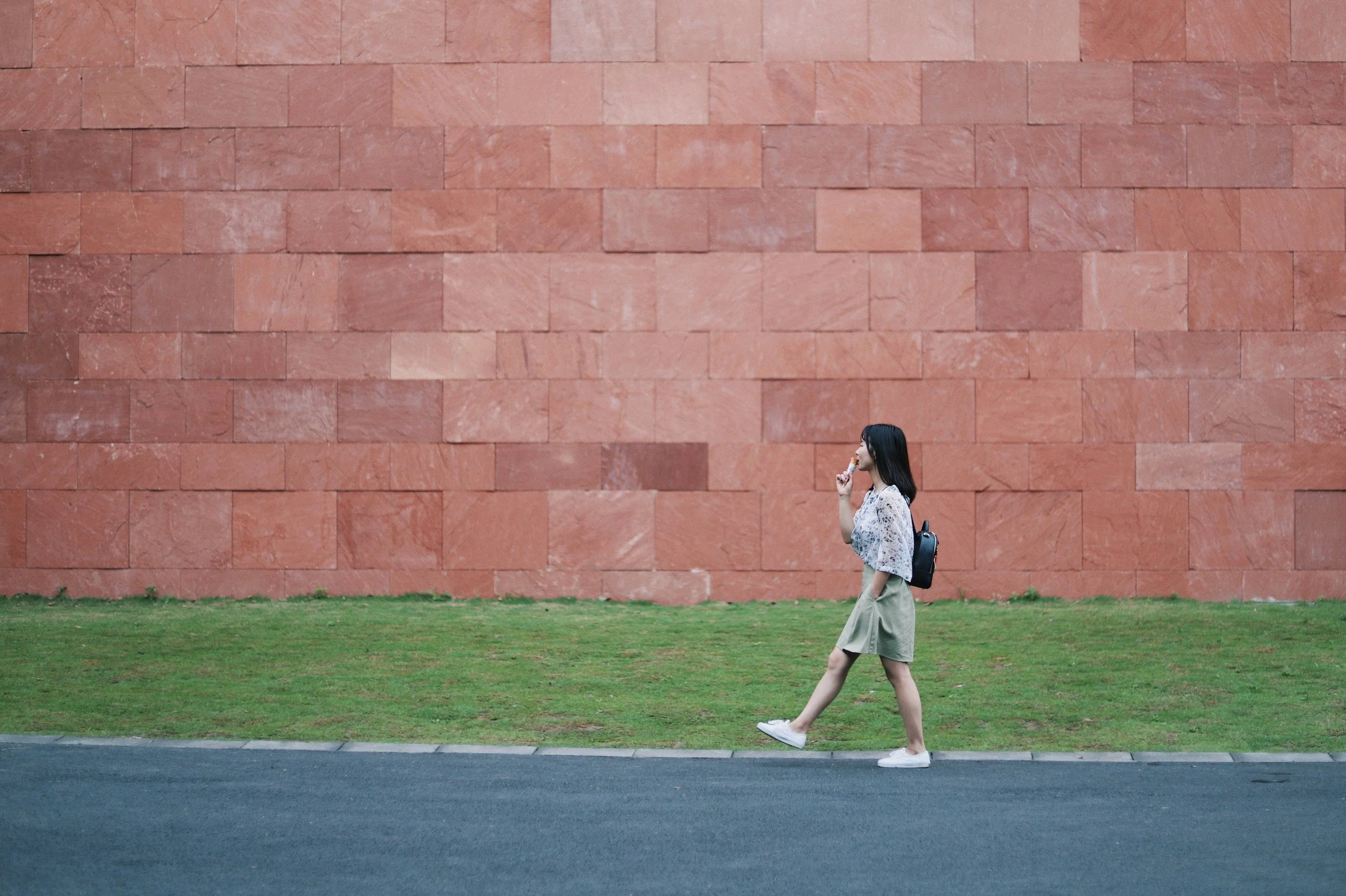 A young woman with black hair, wearing a light-colored lacy top, beige wide-leg pants, white sneakers, and carrying a black backpack, walking on a sidewalk past a large red brick wall and a grassy area.