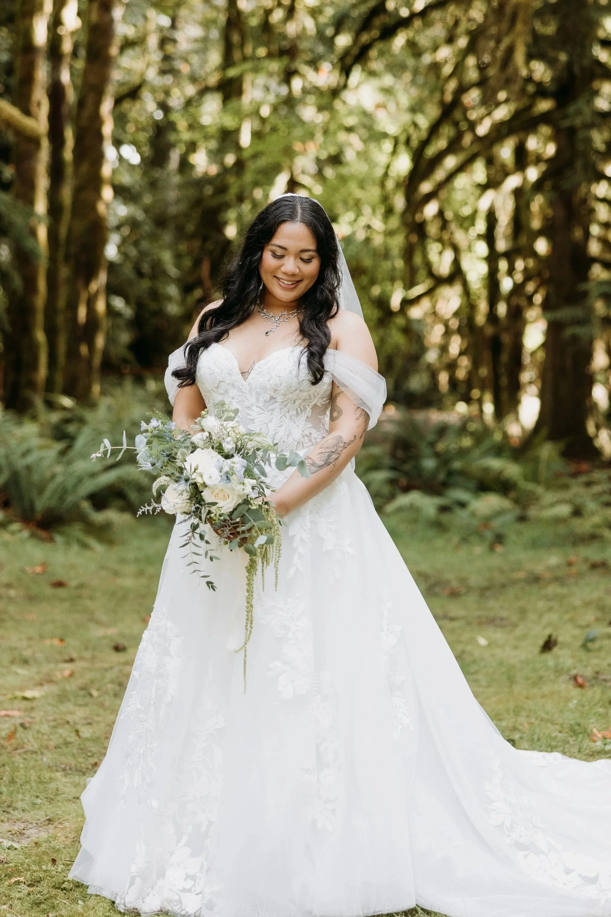 A woman in a white wedding dress holding a bouquet of white and green flowers, standing outdoors in a wooded area, smiling and looking down.