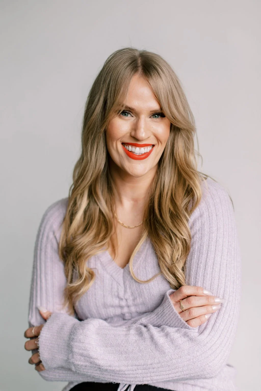 Colleen, wearing red lipstick, smiling, and arms crossed, dressed in a light purple sweater, against a plain grey background.