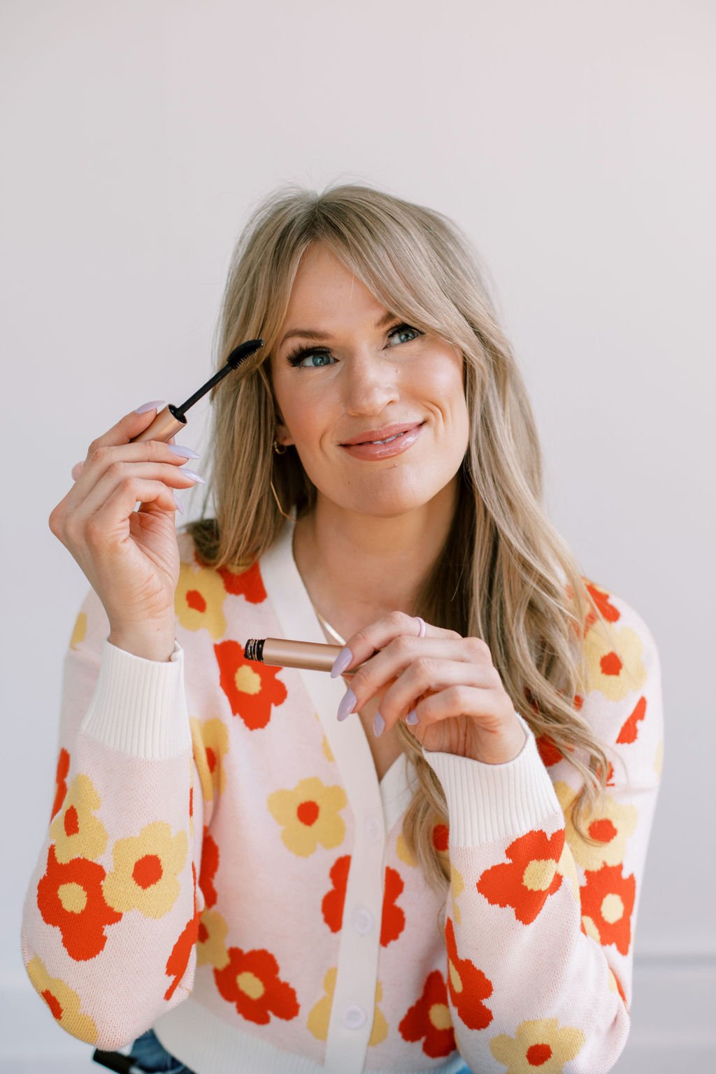 Colleen, wearing a floral sweater, smiling while holding mascara and makeup brush, applying makeup.