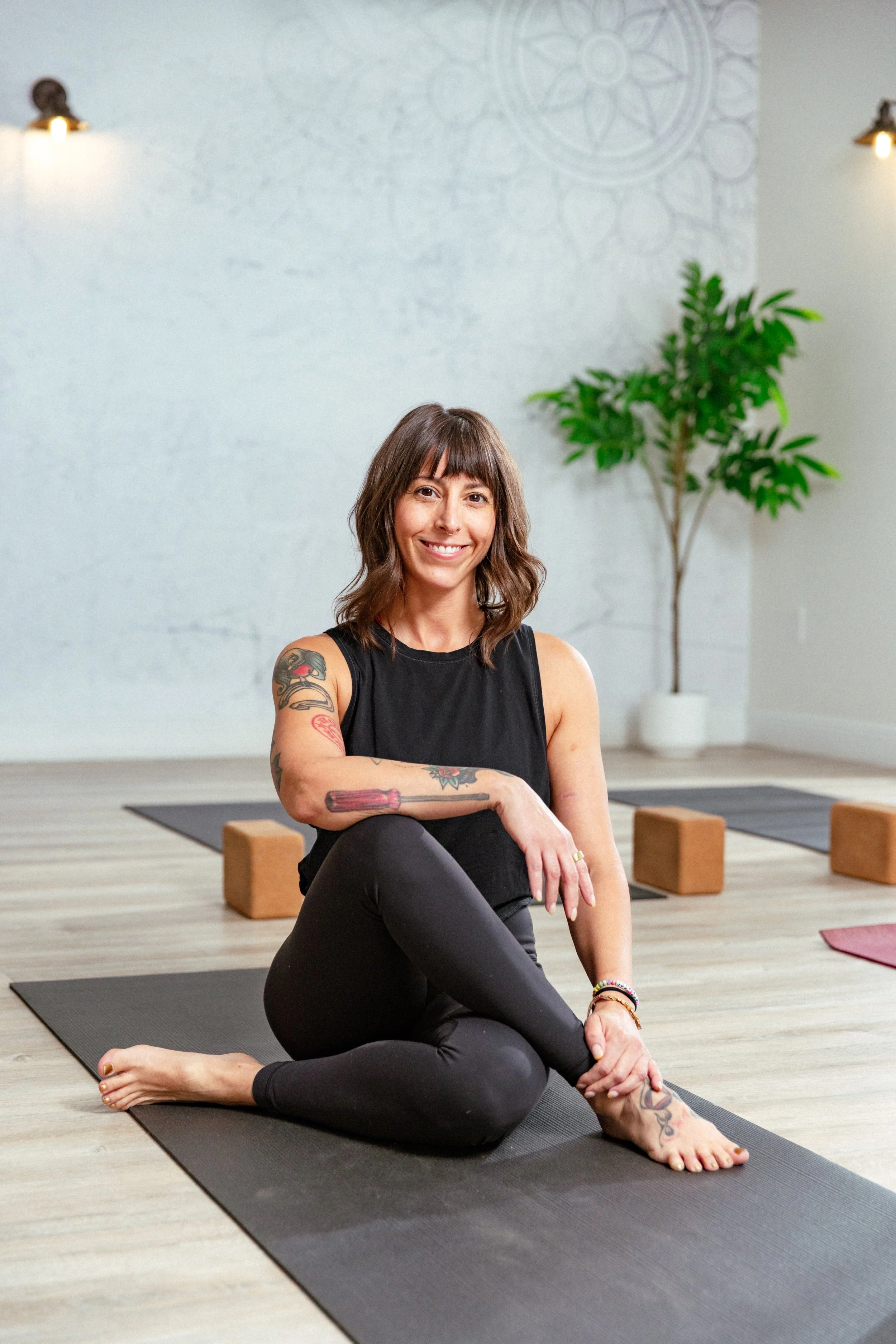 Woman sitting cross-legged on yoga mat in a yoga studio with wooden blocks, green plant, and wall lights.