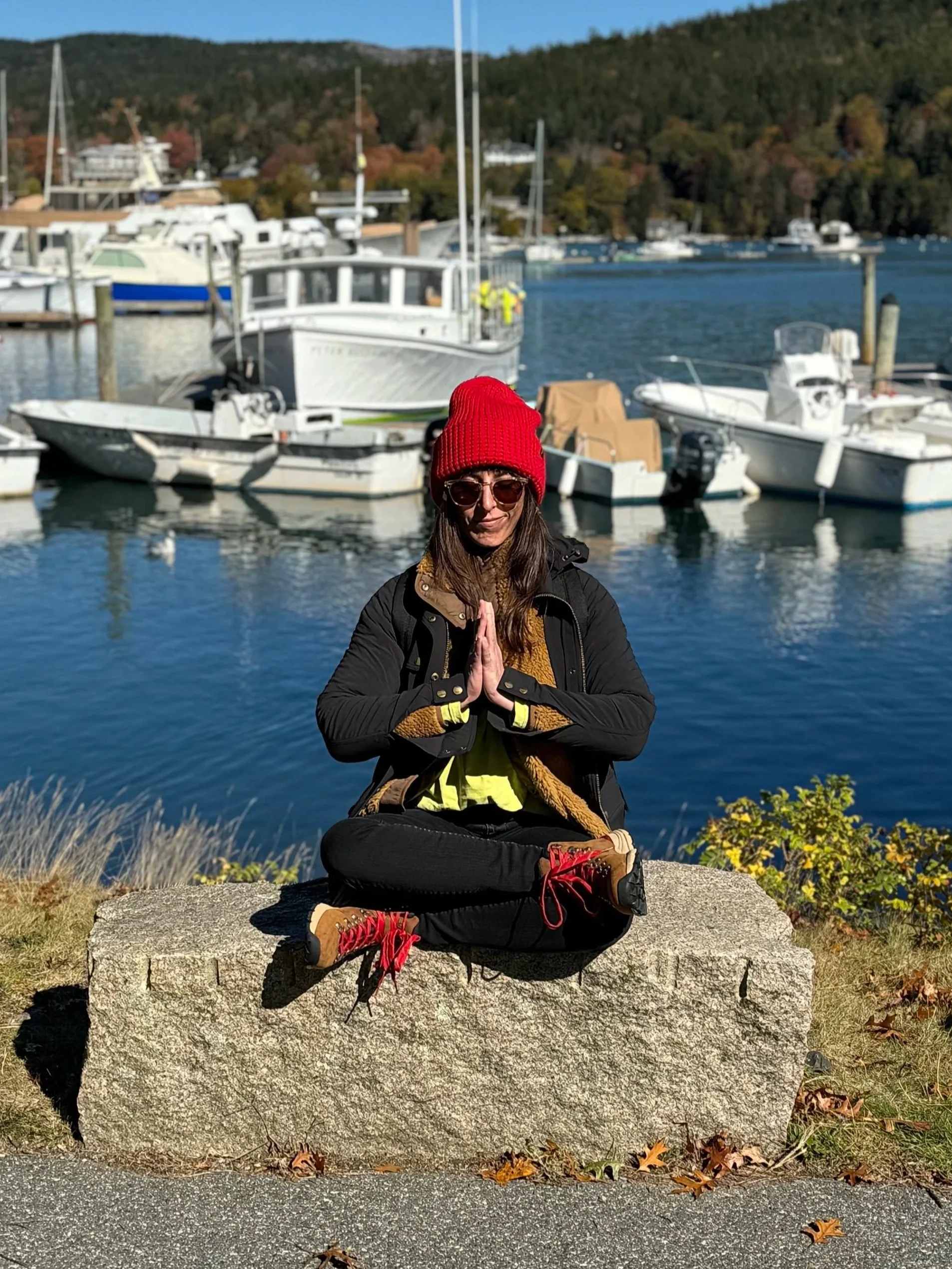Woman practicing meditation or yoga near a harbor with boats, wearing sunglasses, a red beanie, black jacket, and red shoes.
