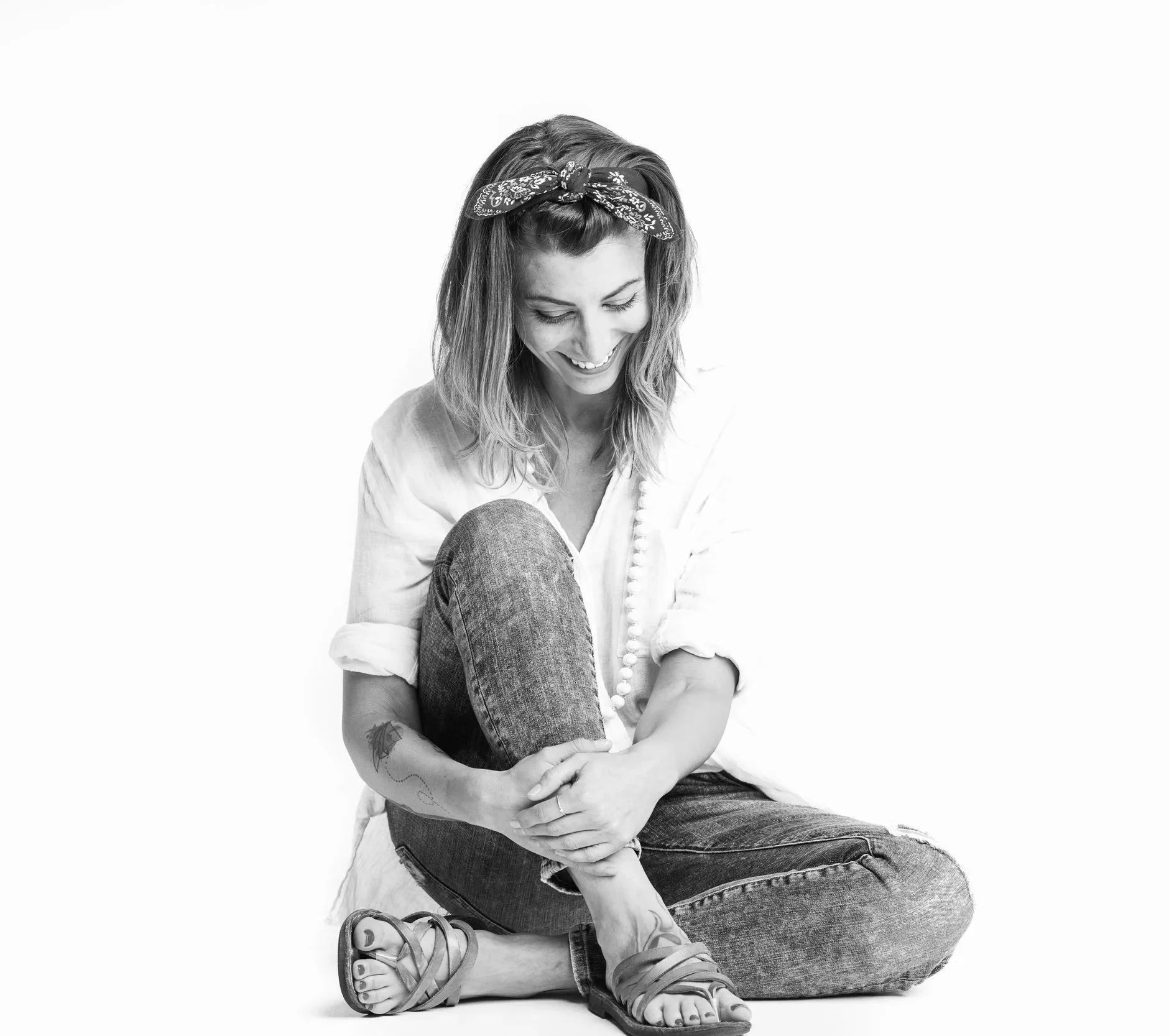 Black and white photo of a woman sitting with her knees drawn up, smiling and looking down, wearing a headscarf and casual clothing.
