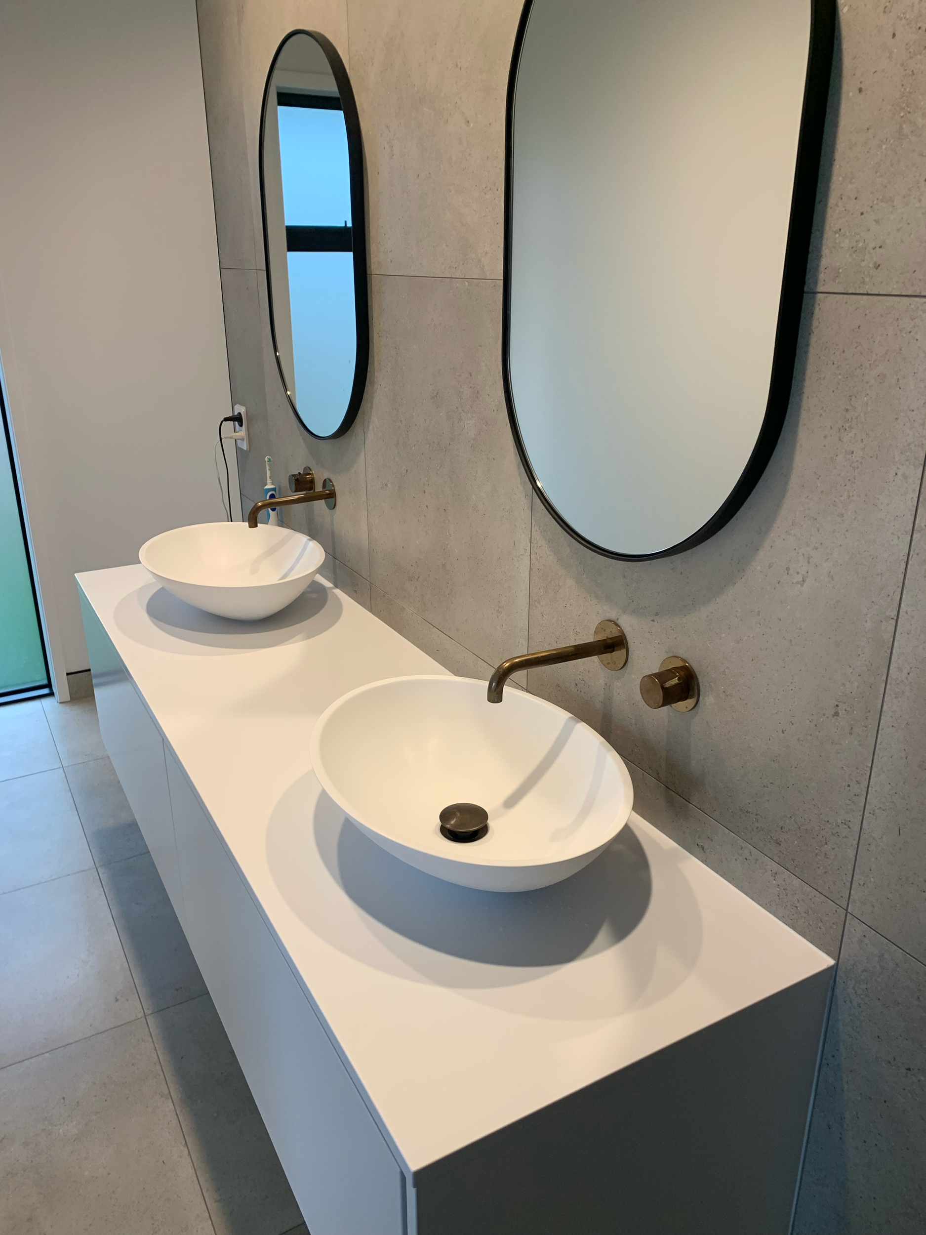 Modern bathroom vanity with two white vessel sinks, brass faucets, large oval mirrors, and a gray countertop.