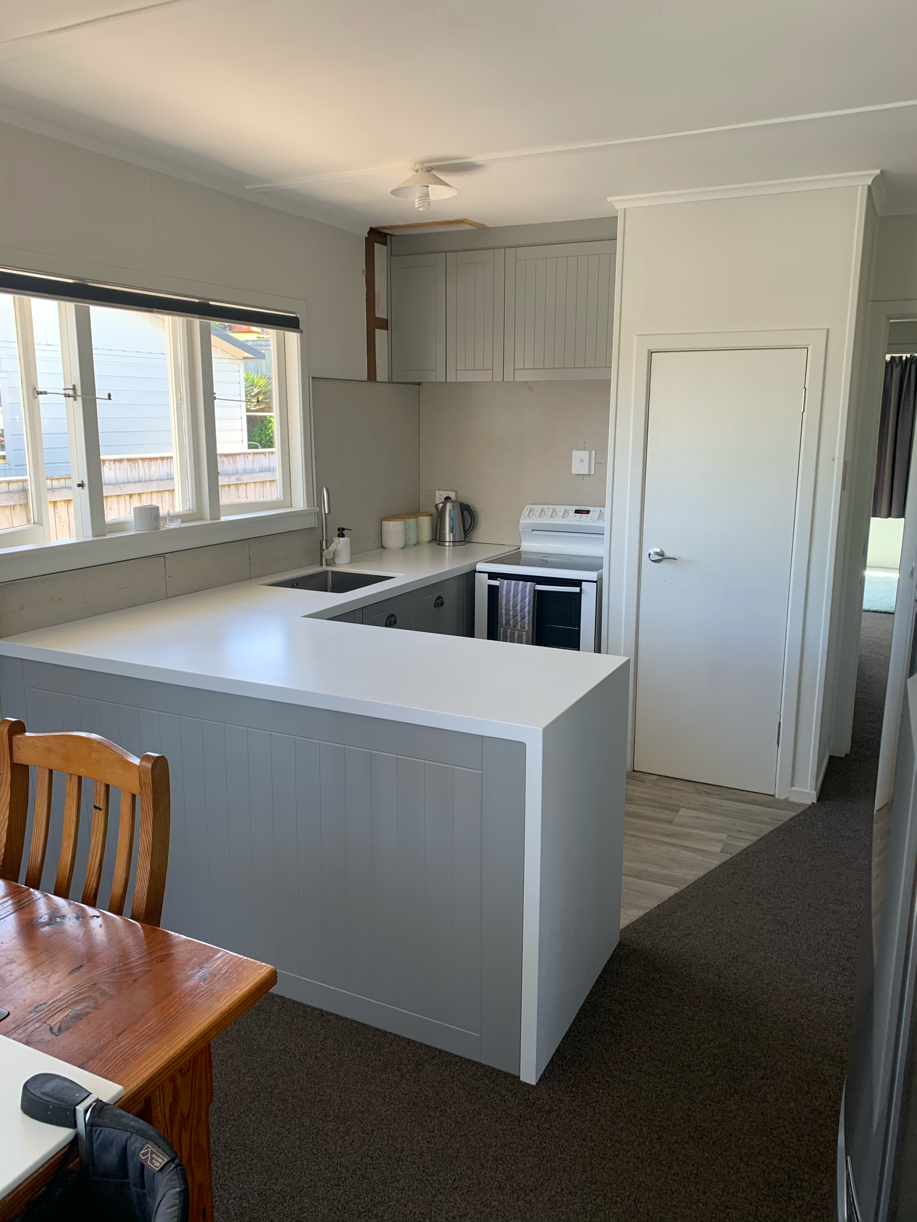 View of a modern kitchen with white cabinets, a white island, a sink, a window, and a wooden dining table with a chair.