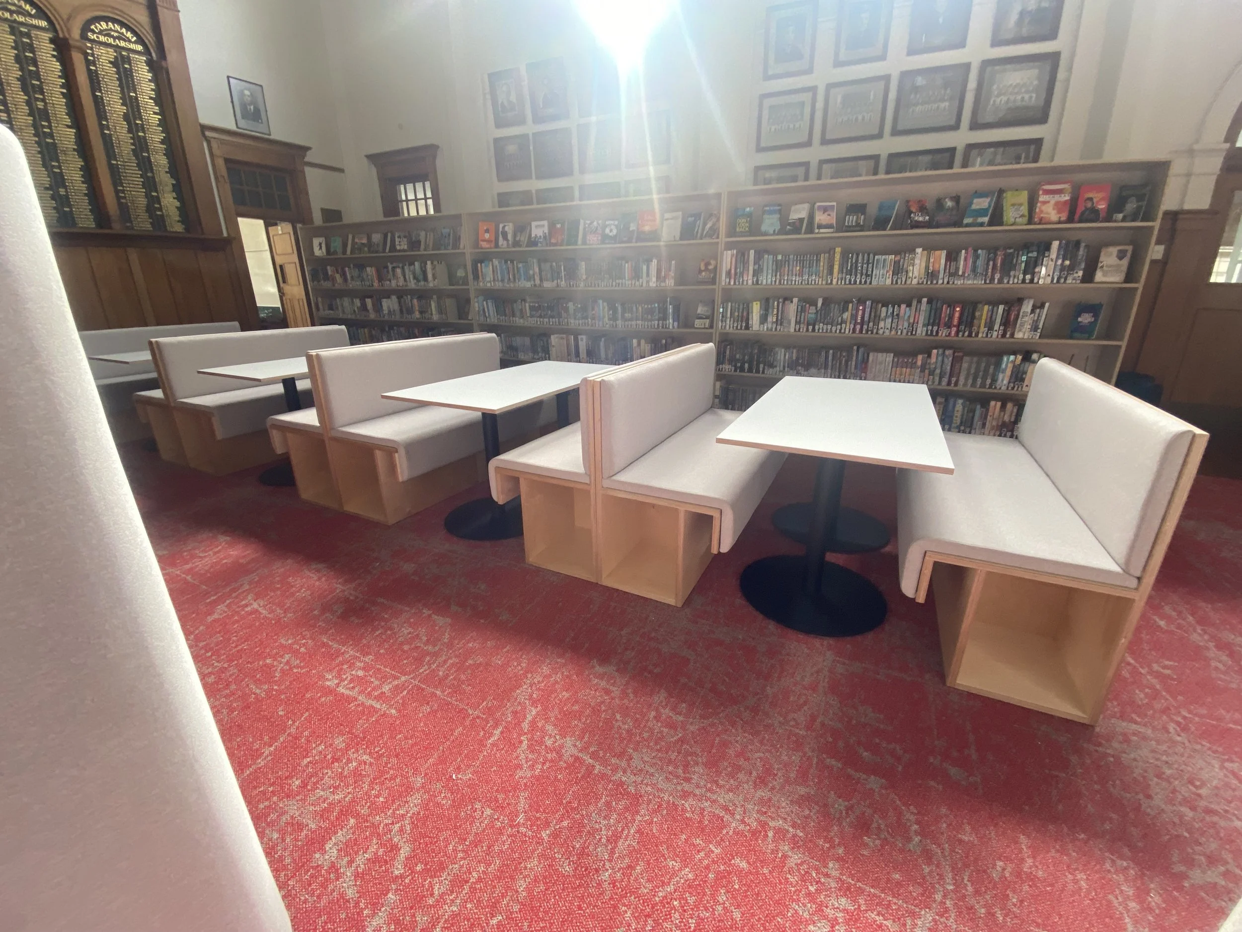 A cozy corner in a library with four white booth seats and white tables, wood-paneled walls, and bookshelves filled with books in the background.