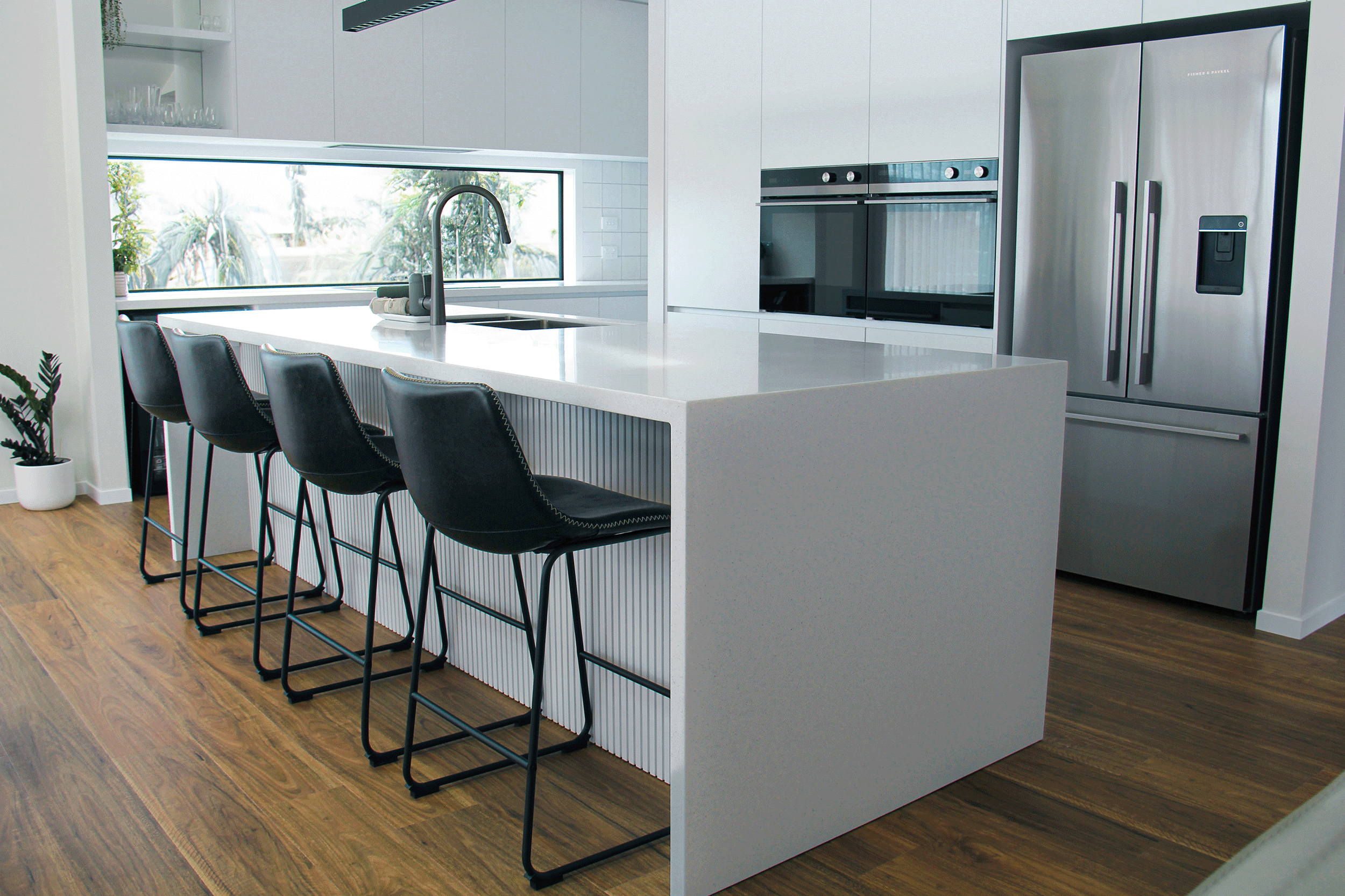 Modern kitchen with white island, four black barstools, stainless steel refrigerator, built-in oven, and a horizontal window showing greenery outside.