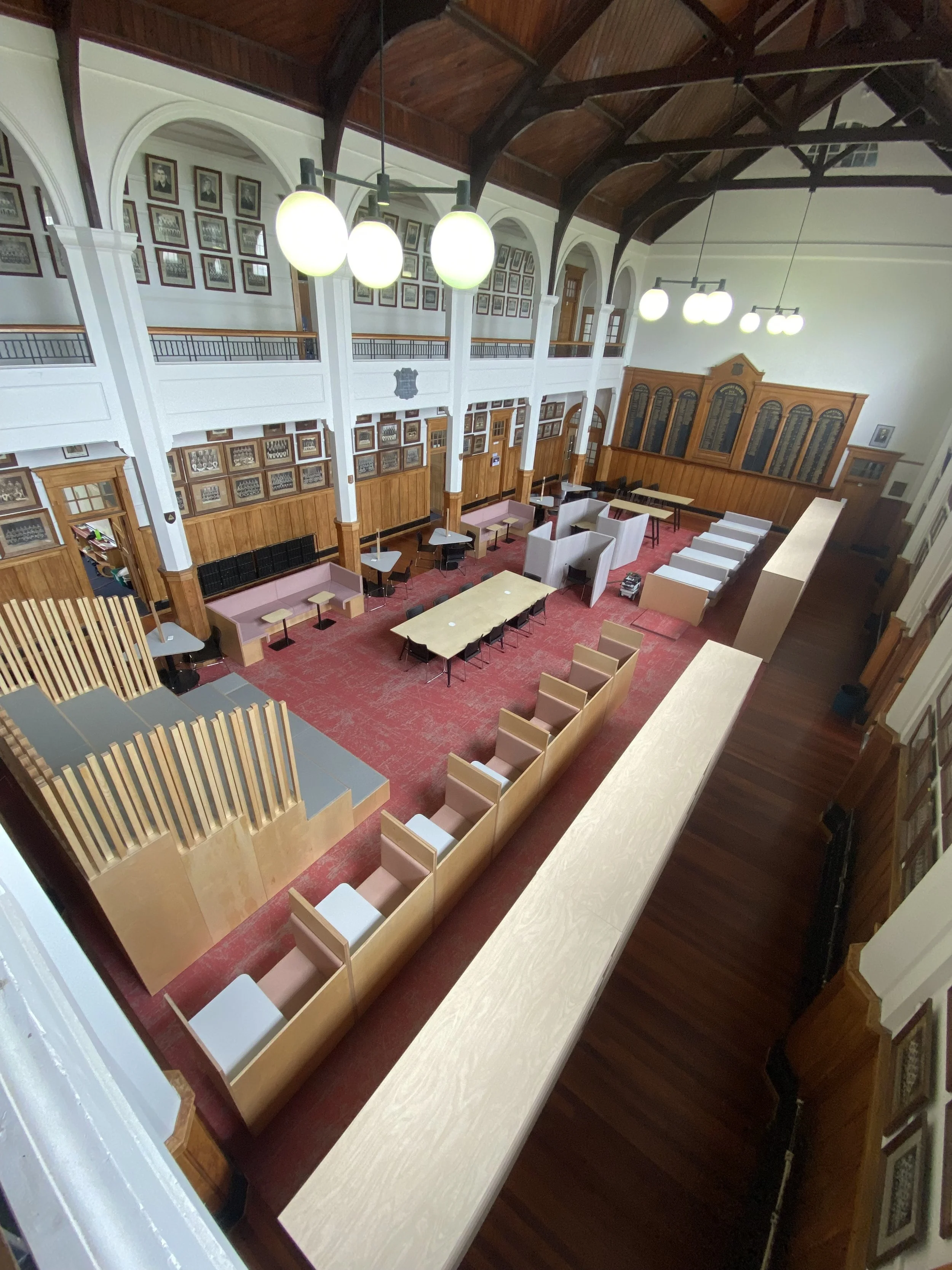 An interior view of a large, historic room with a high arched wooden ceiling, hanging spherical light fixtures, and wood-paneled walls decorated with framed pictures. The room has various seating arrangements including chairs, sofas, and tables, with a red carpet covering the floor.