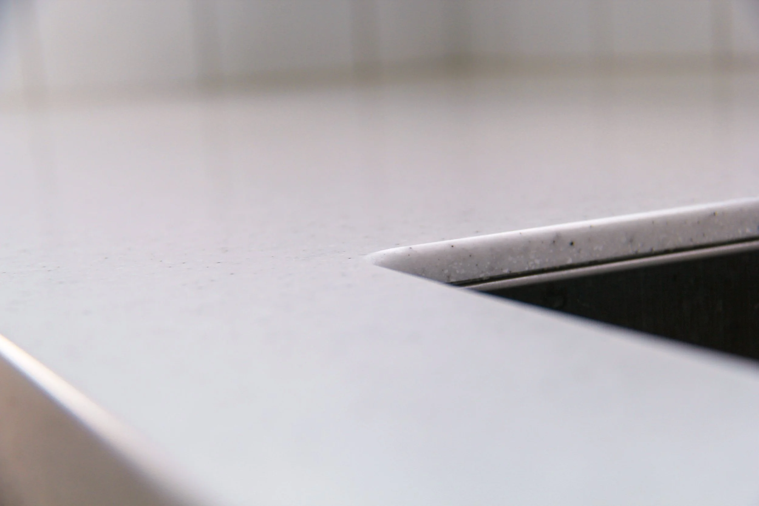 A close-up of a smooth, speckled white kitchen countertop with a stainless steel undermounted sink. The image emphasizes the clean, modern surface and edging details.