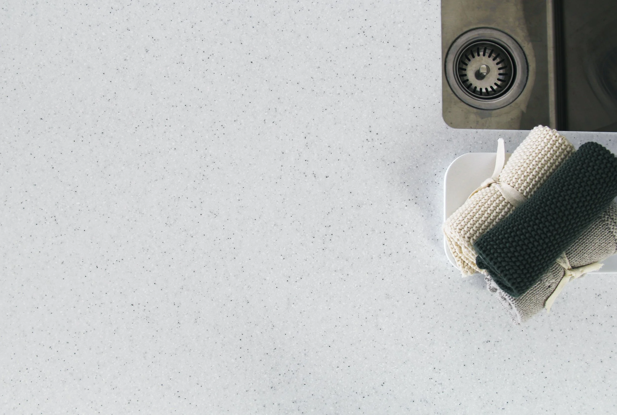 Two rolled-up knit towels, one beige and one dark green, placed on a white dish drainer on a speckled white countertop near a sink.