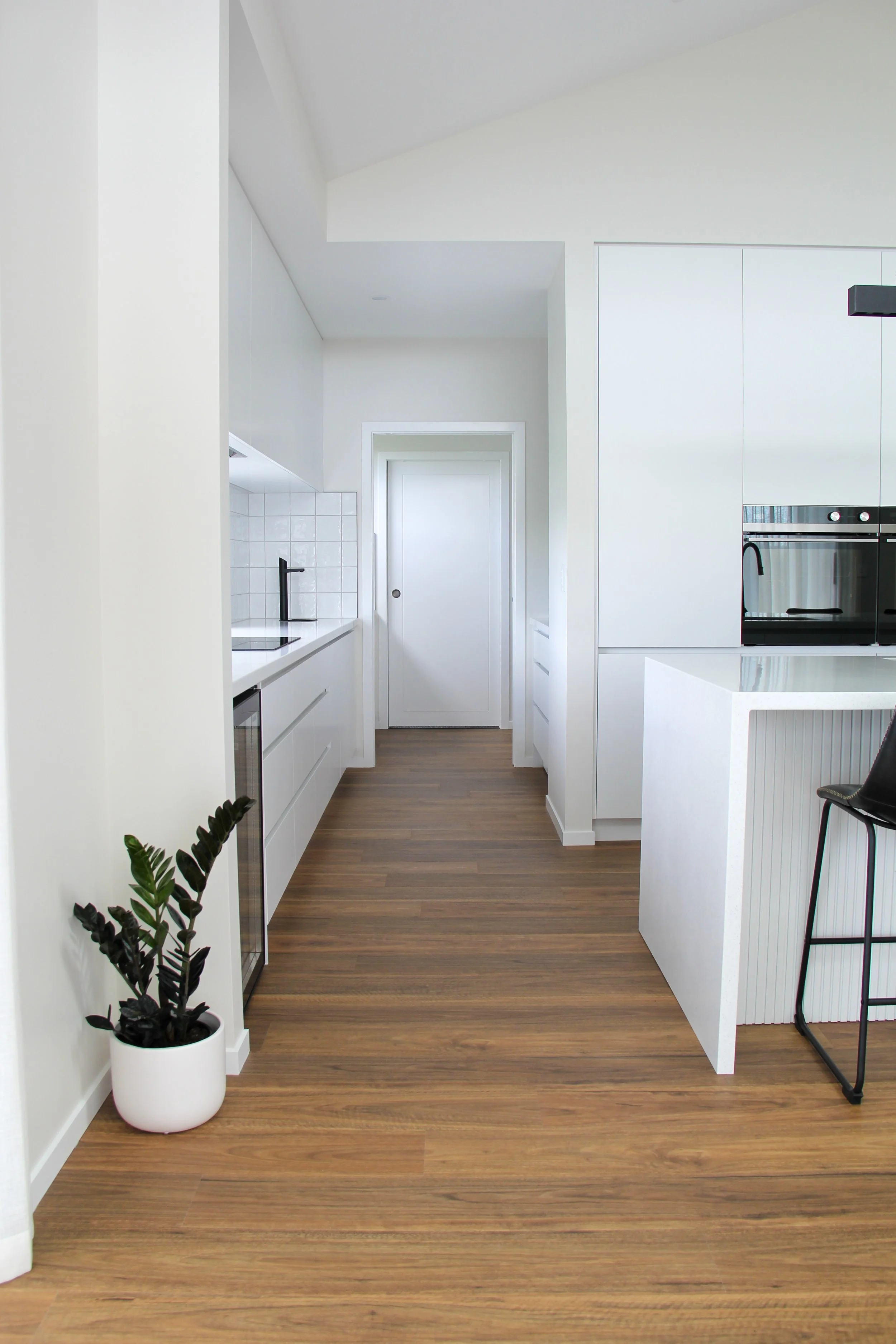 Modern kitchen with white cabinets, a black faucet, a small wine fridge, wooden flooring, a black chair, and a potted plant, seen from a hallway