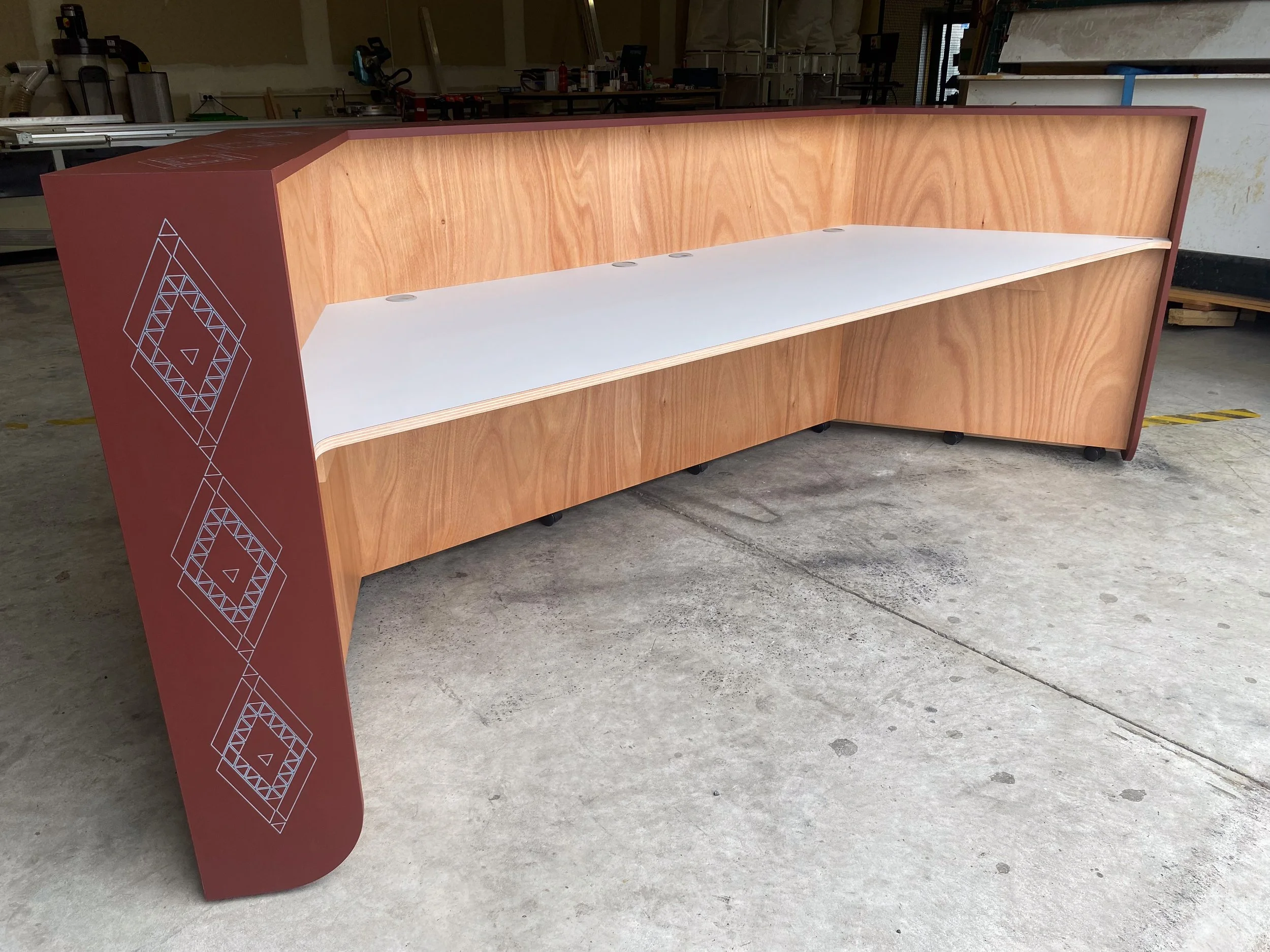 Wooden reception or conference desk with a decorative geometric pattern on the side, situated in a workshop or storage area.