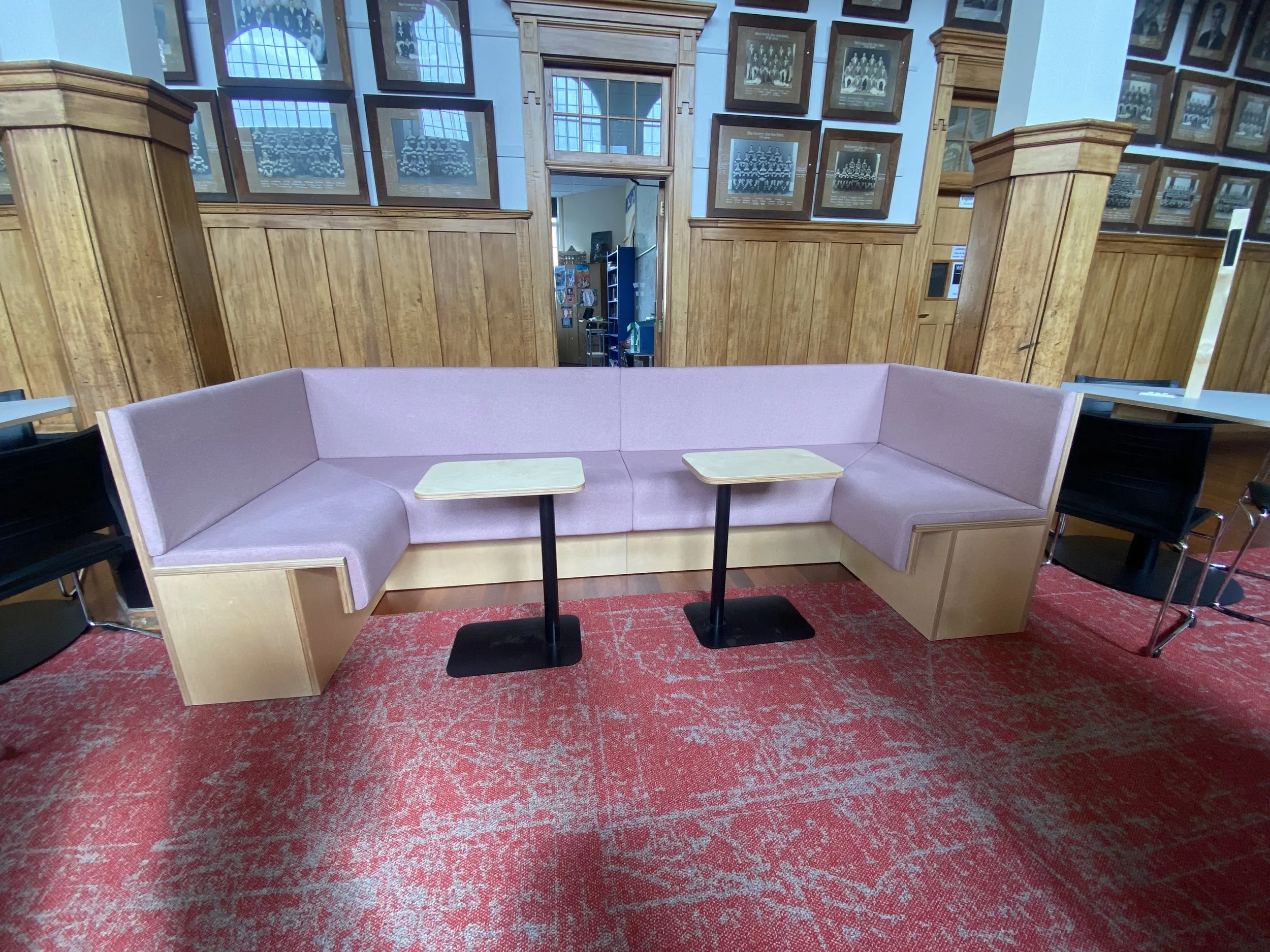 Pink upholstered corner seating with wooden bases and two small tables in a room with wood-paneled walls and framed photographs.