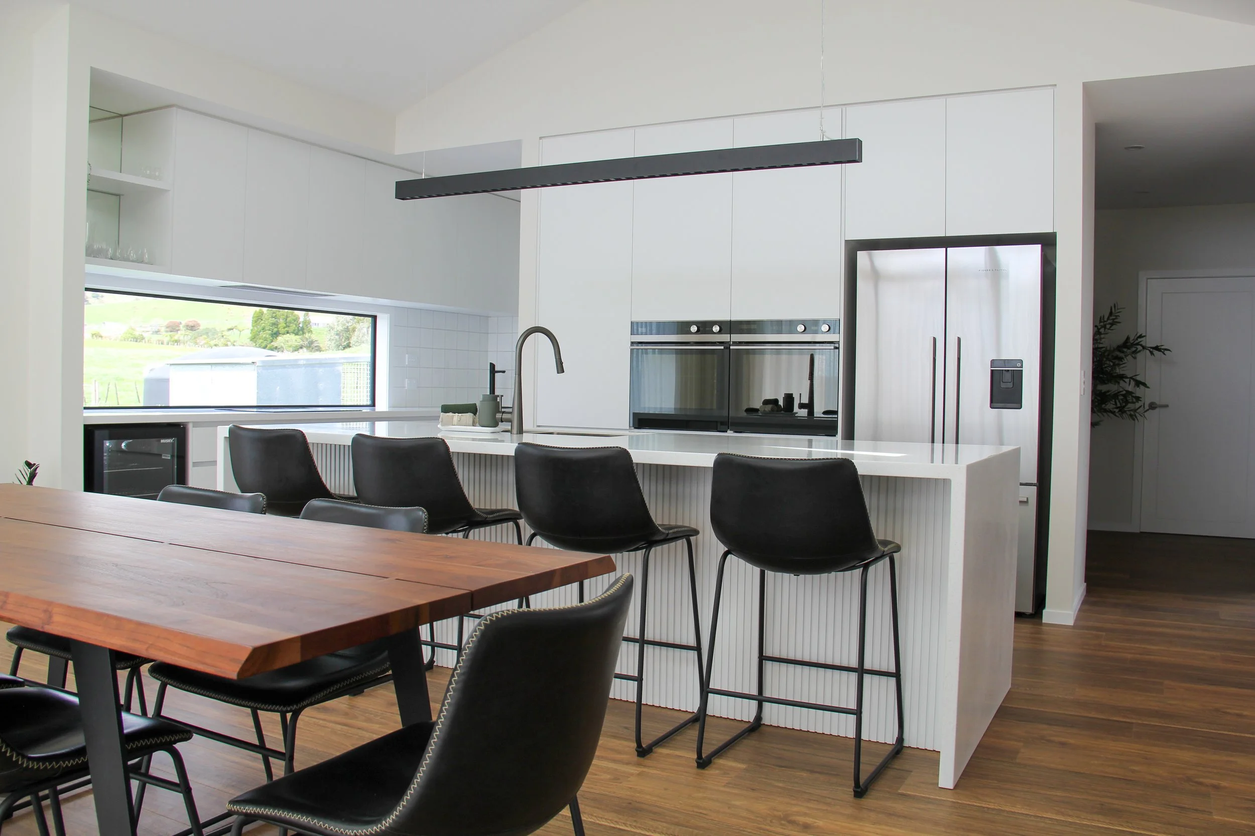 Modern kitchen with white cabinetry, stainless steel appliances, a black hanging light fixture, black bar stools at a white island, and a wooden dining table with black chairs. Large window overlooks a green landscape.