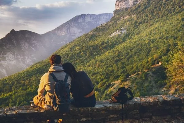 Couple sitting on a scenic overlook facing a majestic mountain range under a clear sky.