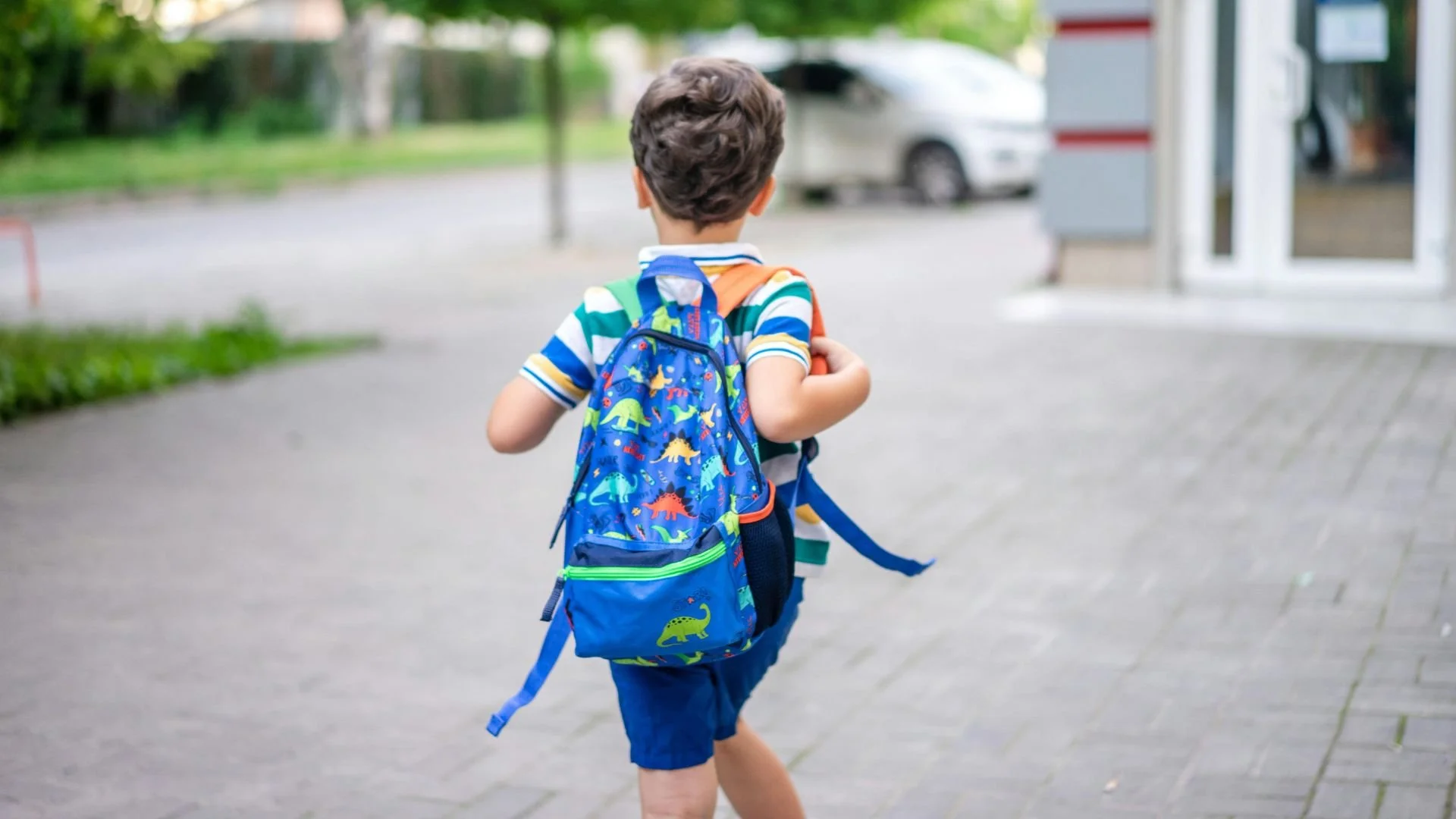 Back view of a young boy carrying a blue dinosaur backpack while walking down a sidewalk toward school.