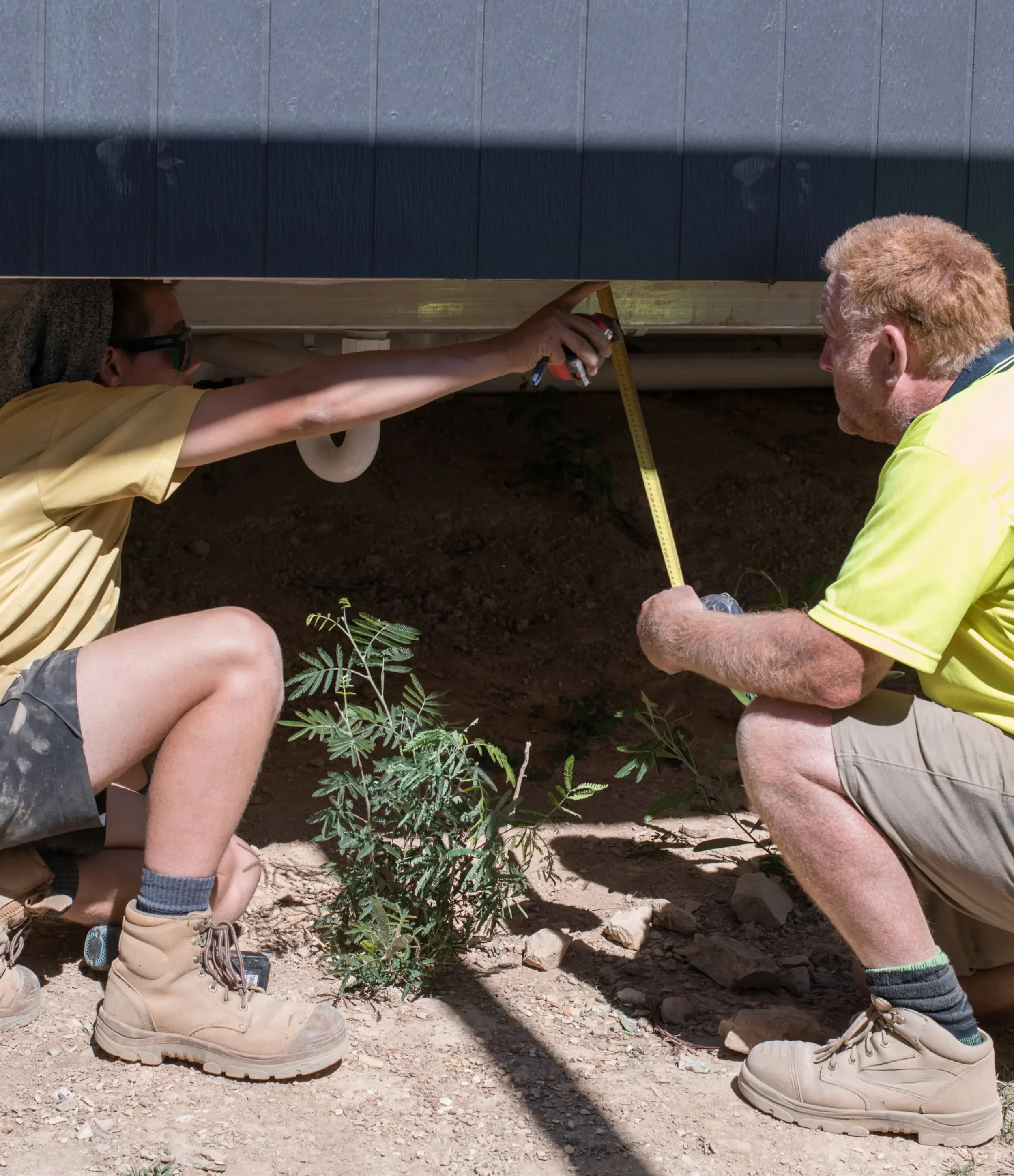 Two people using a tape measure to measure up underneath a house