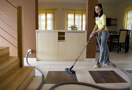 A woman vacuuming a carpet in a brightly lit living room with yellow walls and windows.