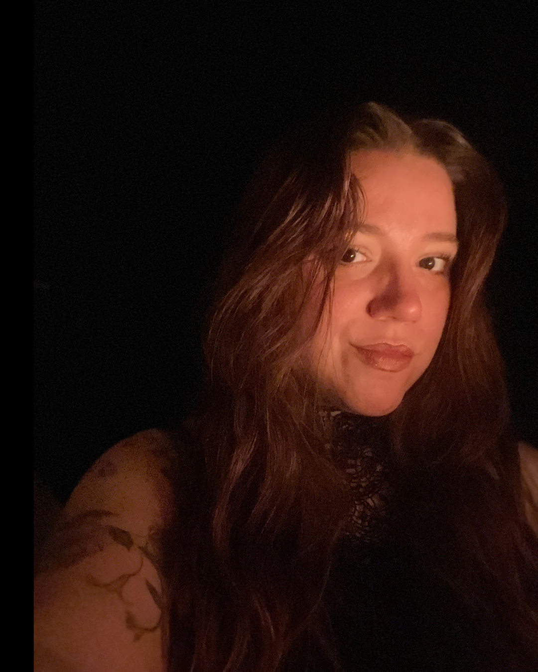 A woman with long wavy brown hair and a tattoo on her arm, taking a selfie in low lighting with a dark background.