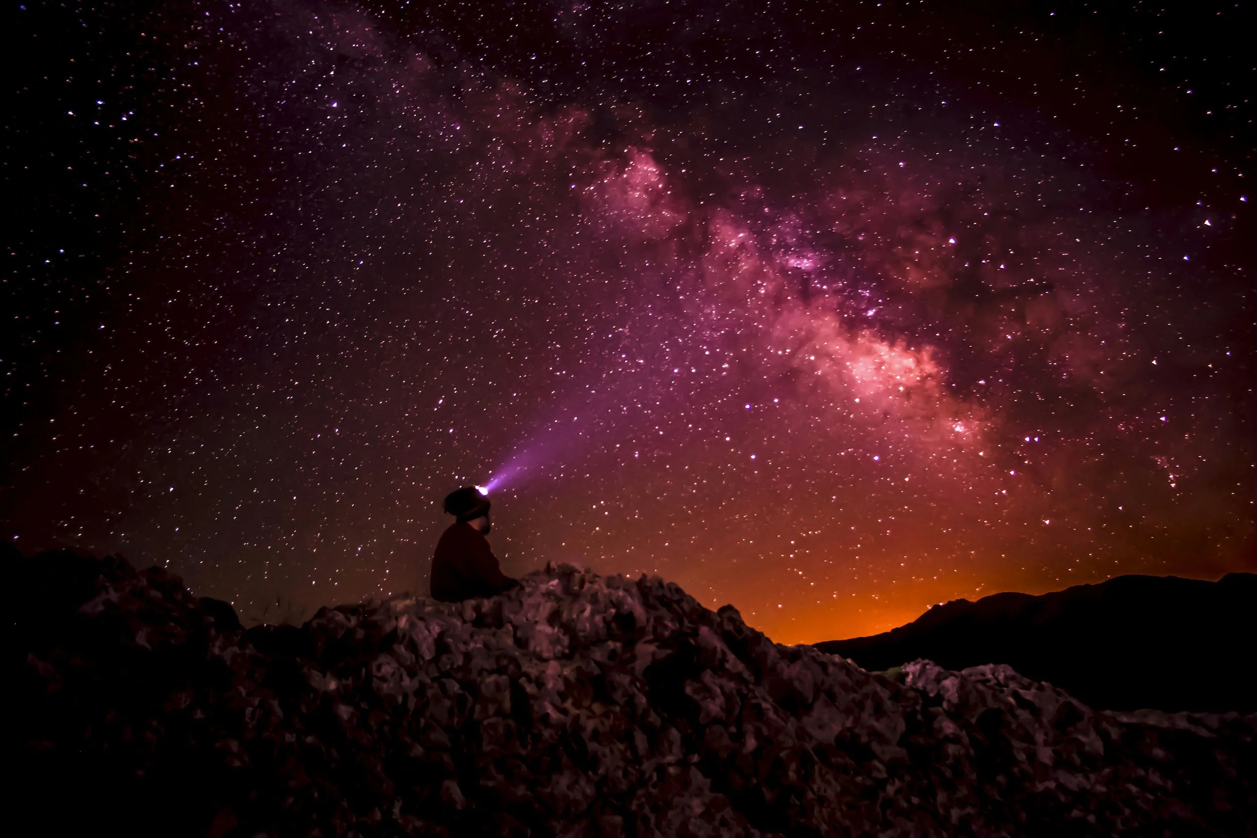 Person sitting on rocks watching the starry night sky with the Milky Way galaxy, while wearing a headlamp that shines purple light.