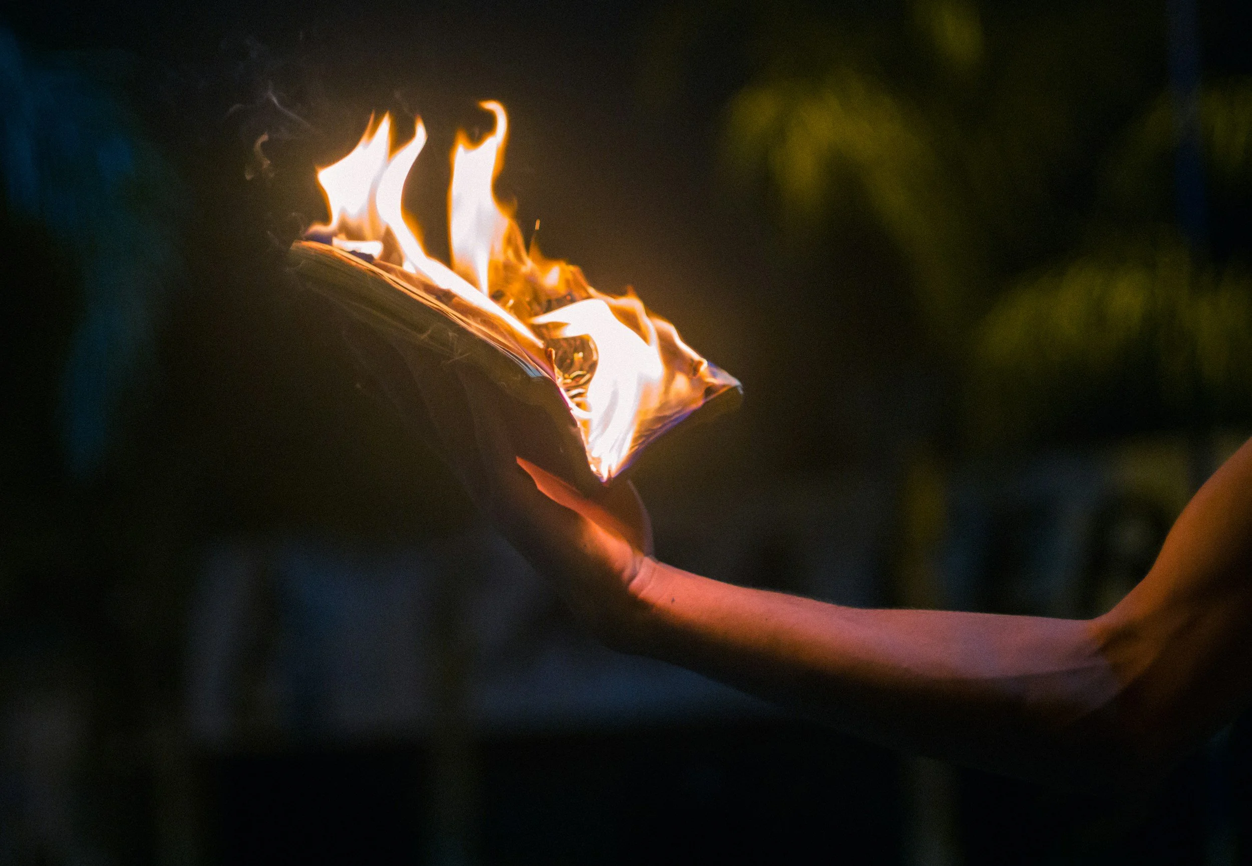 A hand holding a small object, possibly a book or a piece of paper, which is on fire with bright flames at night or in a dark setting.