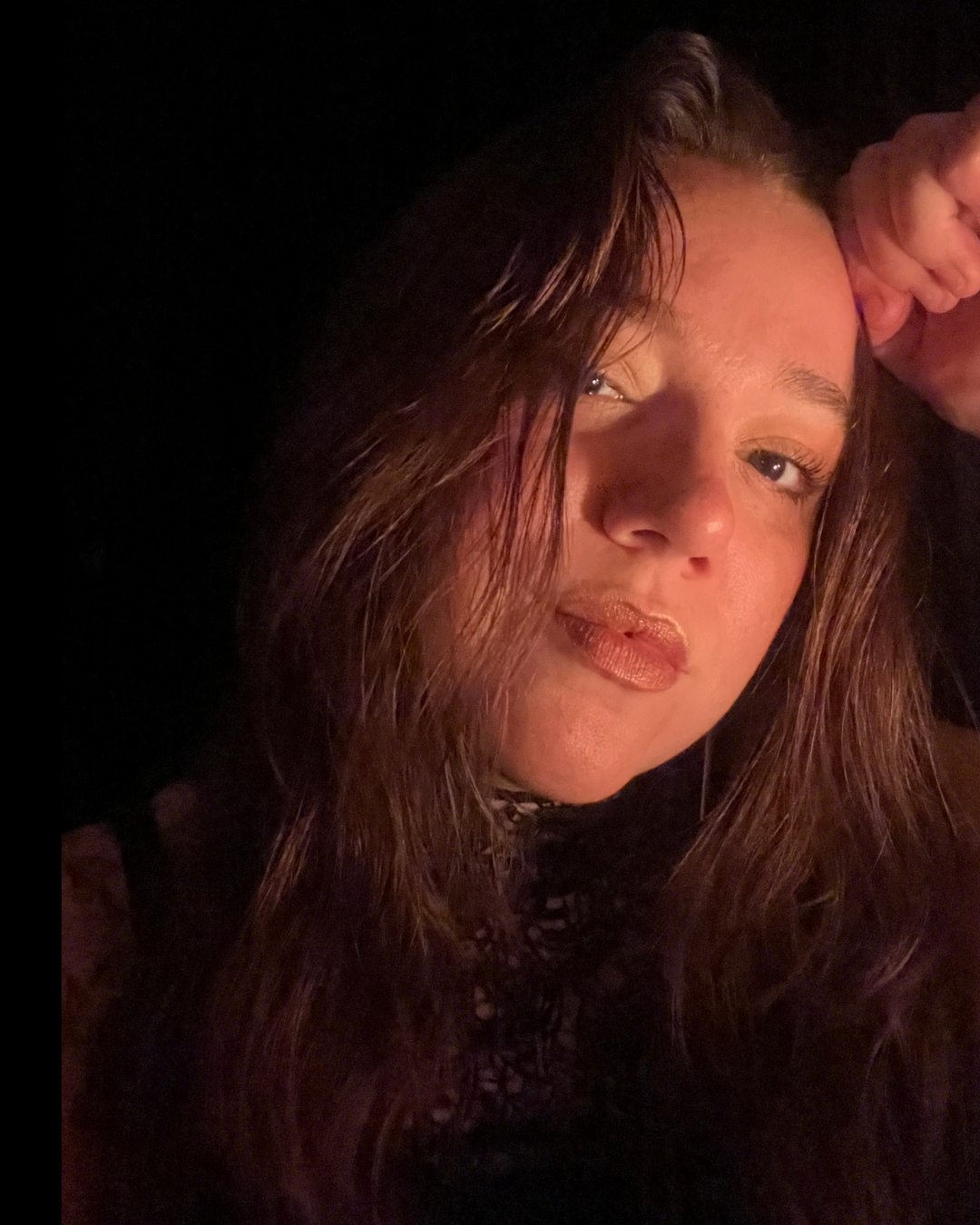 A close-up photo of a young woman with long, wavy brown hair, light skin with freckles, wearing makeup including lipstick, and looking at the camera while lying down in a dimly lit environment.