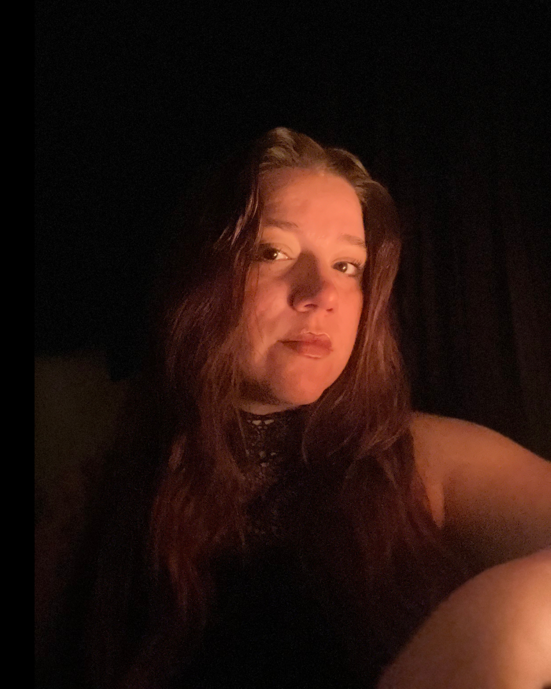 A woman with long, wavy red hair is looking at the camera in a dark setting with soft lighting.