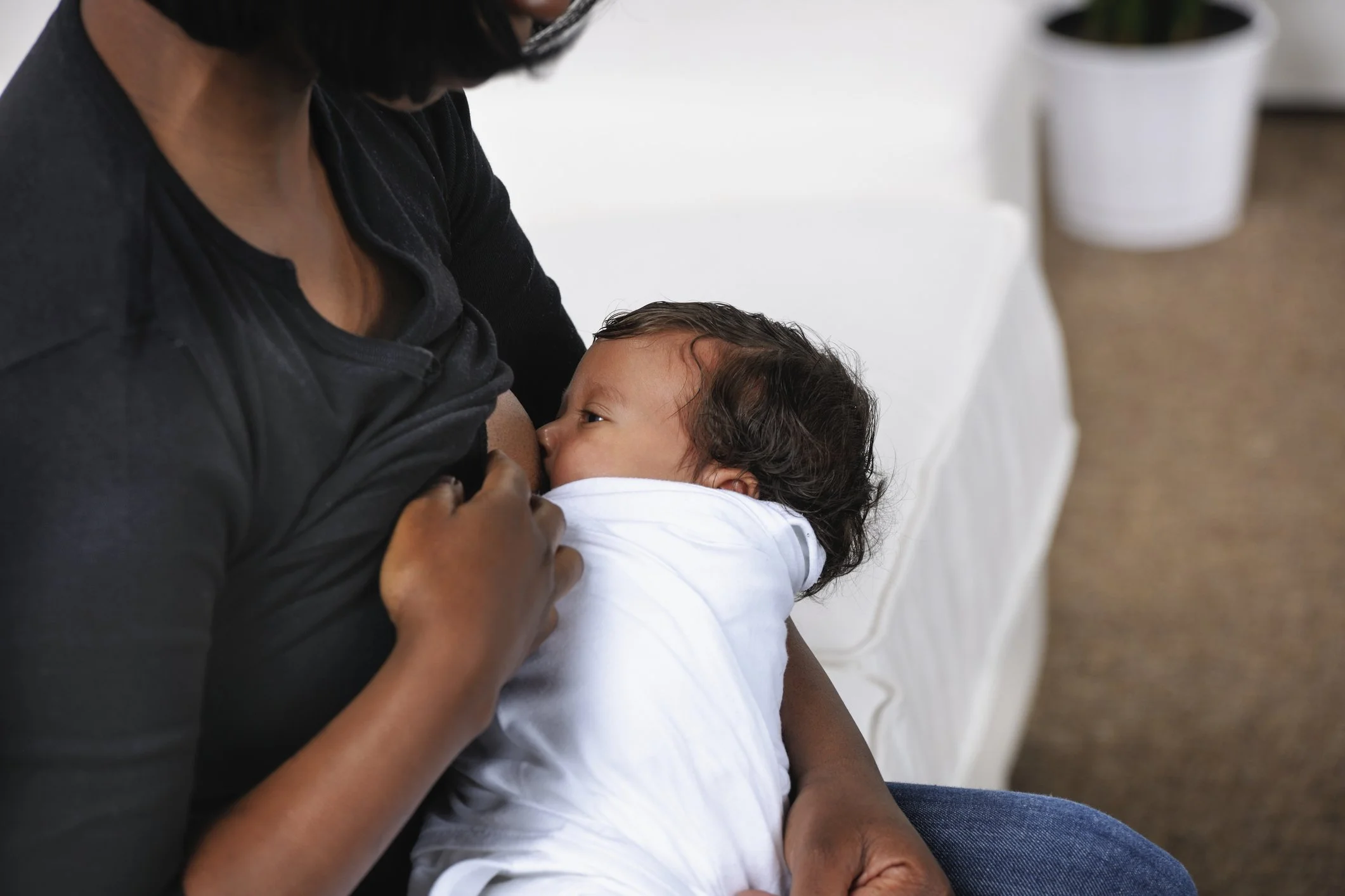 A person breastfeeding a baby while sitting on a white surface, with a white pot plant in the background.