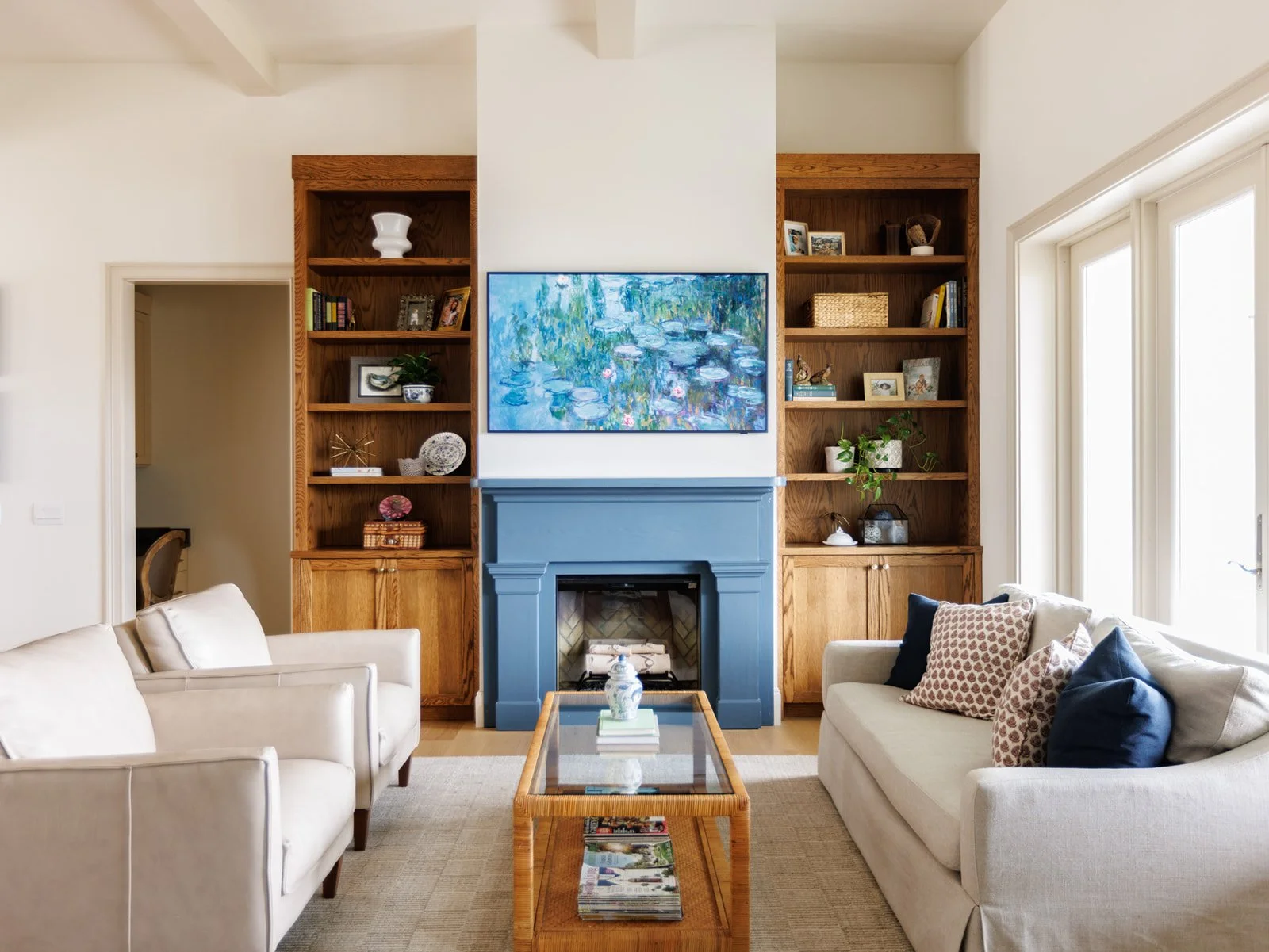 Living room with beige sofas, a fireplace with a blue surround, a wall-mounted painting of water lilies, built-in wooden bookshelves on either side of the fireplace with decorative items and books, and large windows allowing natural light.