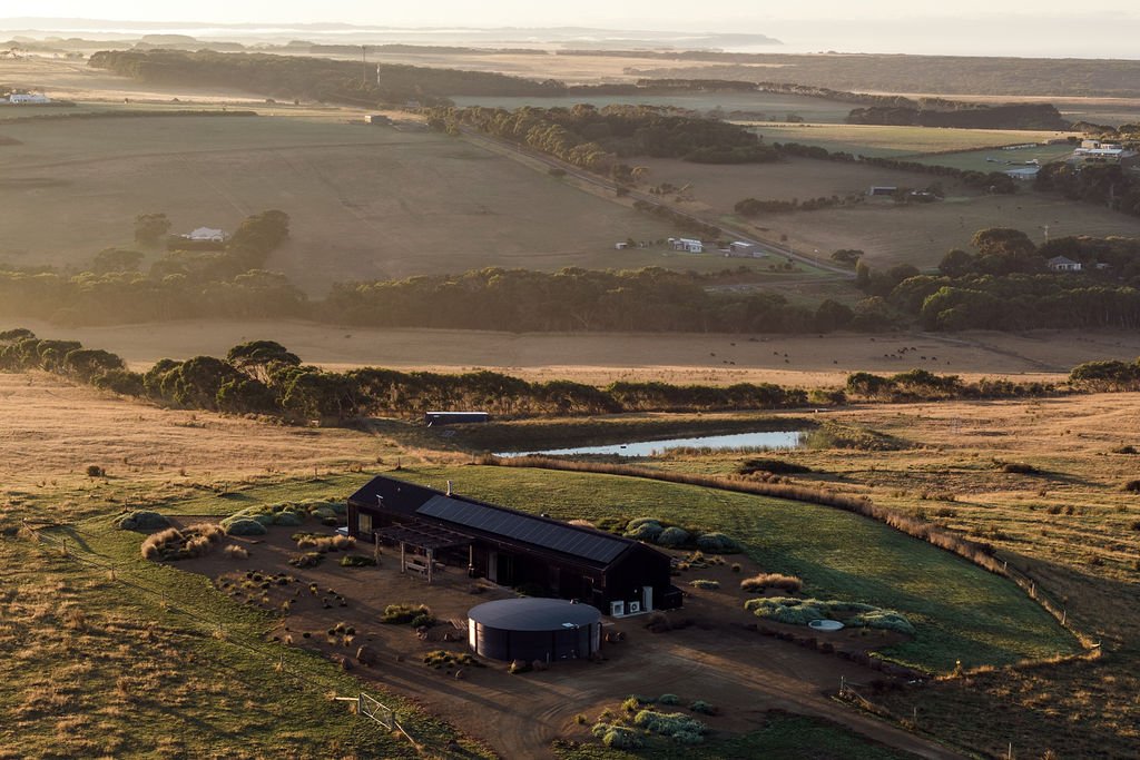 Aerial image showing Port Farm - a black shed like house with water tank and coastal garden. The view takes in farm dams and paddocks.