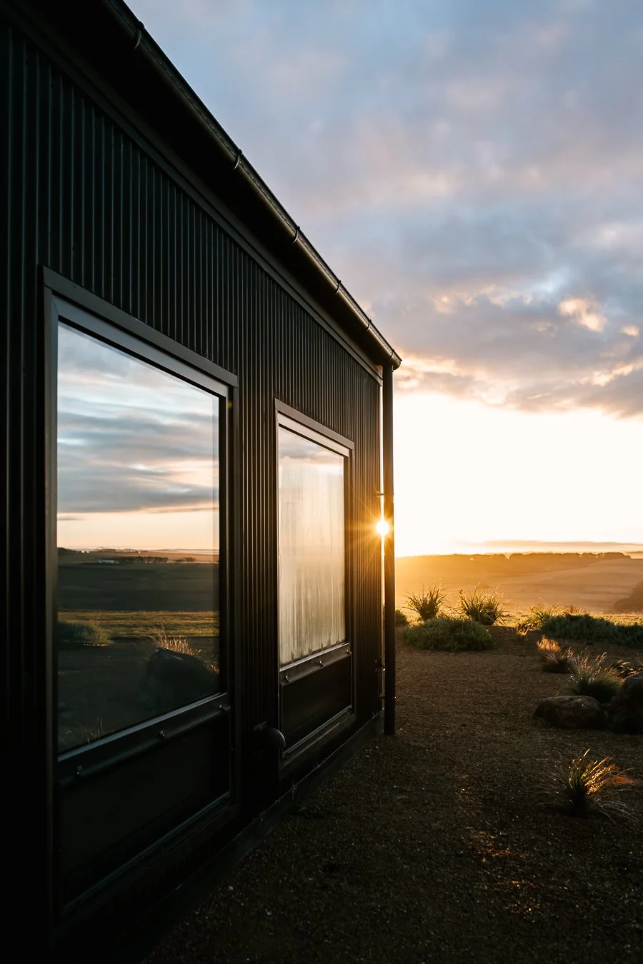A black building with large windows reflecting the landscape at sunset, with the sun partially behind the building, and a scenic view of open land and cloudy sky in the background.