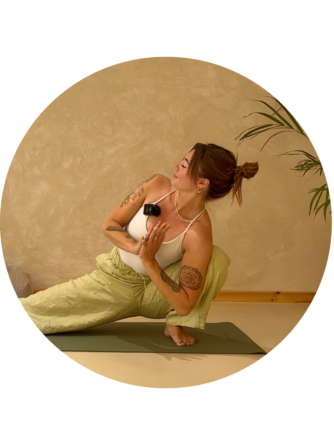 A woman practicing yoga indoors on a mat, performing a side bend stretch with one hand on her chest and the other reaching upward, with tattoos on her arms and a plant visible in the background.