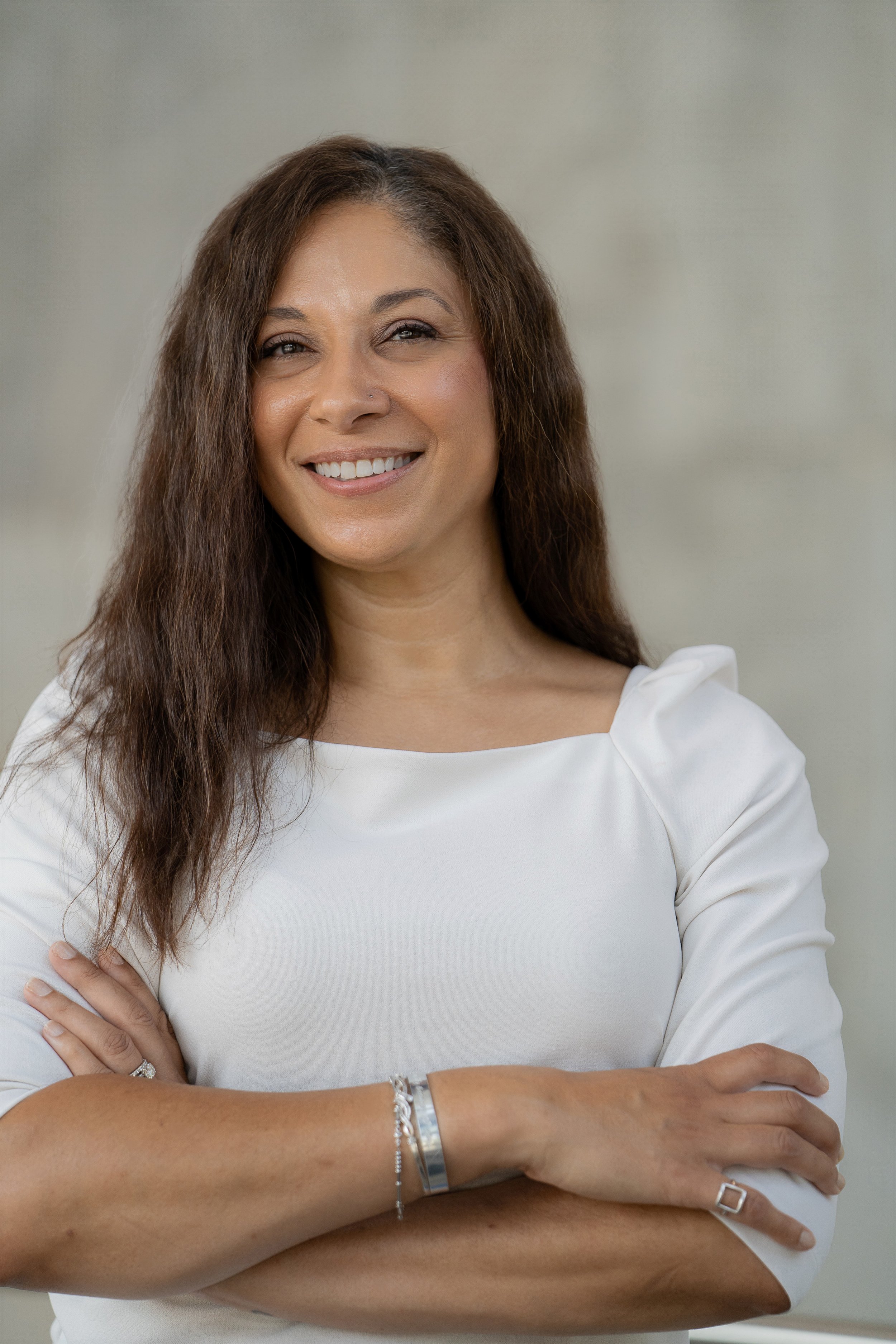 Portrait of a woman smiling with crossed arms, wearing a white top and jewelry, standing against a neutral background.