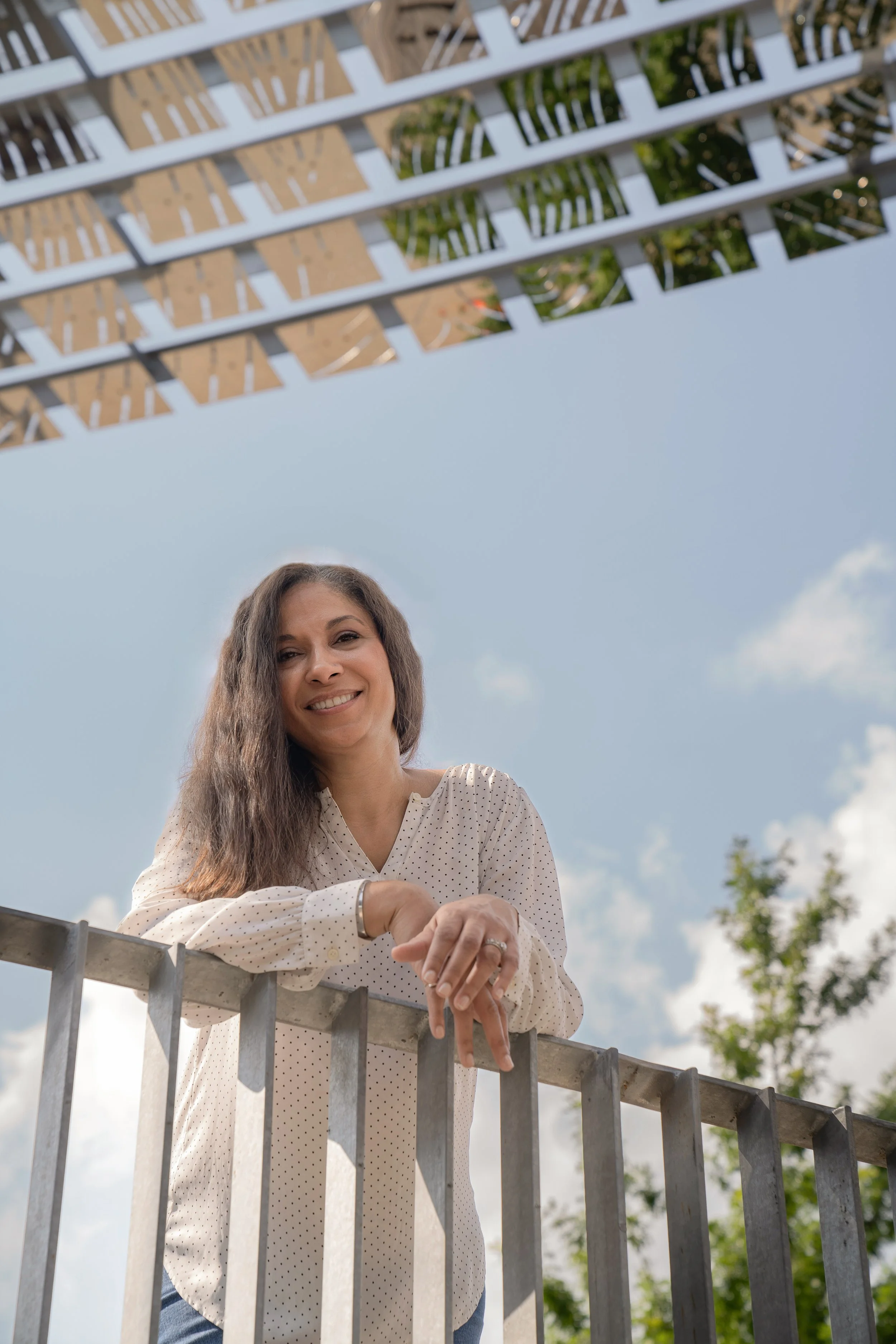 Smiling woman with long brown hair leaning on metal railing outdoors, blue sky with some clouds and a tree in the background.