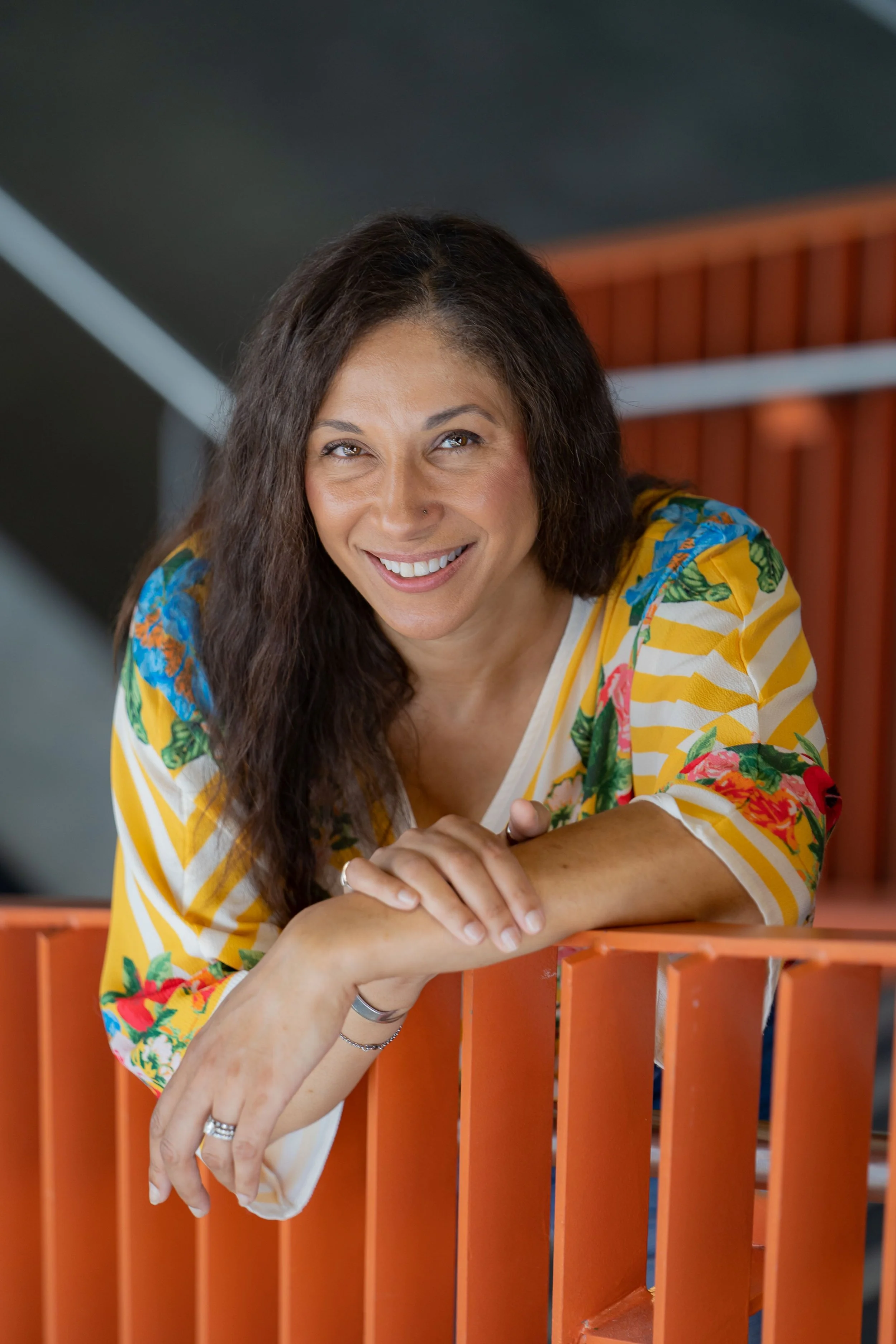 A woman with dark wavy hair, smiling, leaning on an orange railing, wearing a colorful, floral and striped yellow shirt.