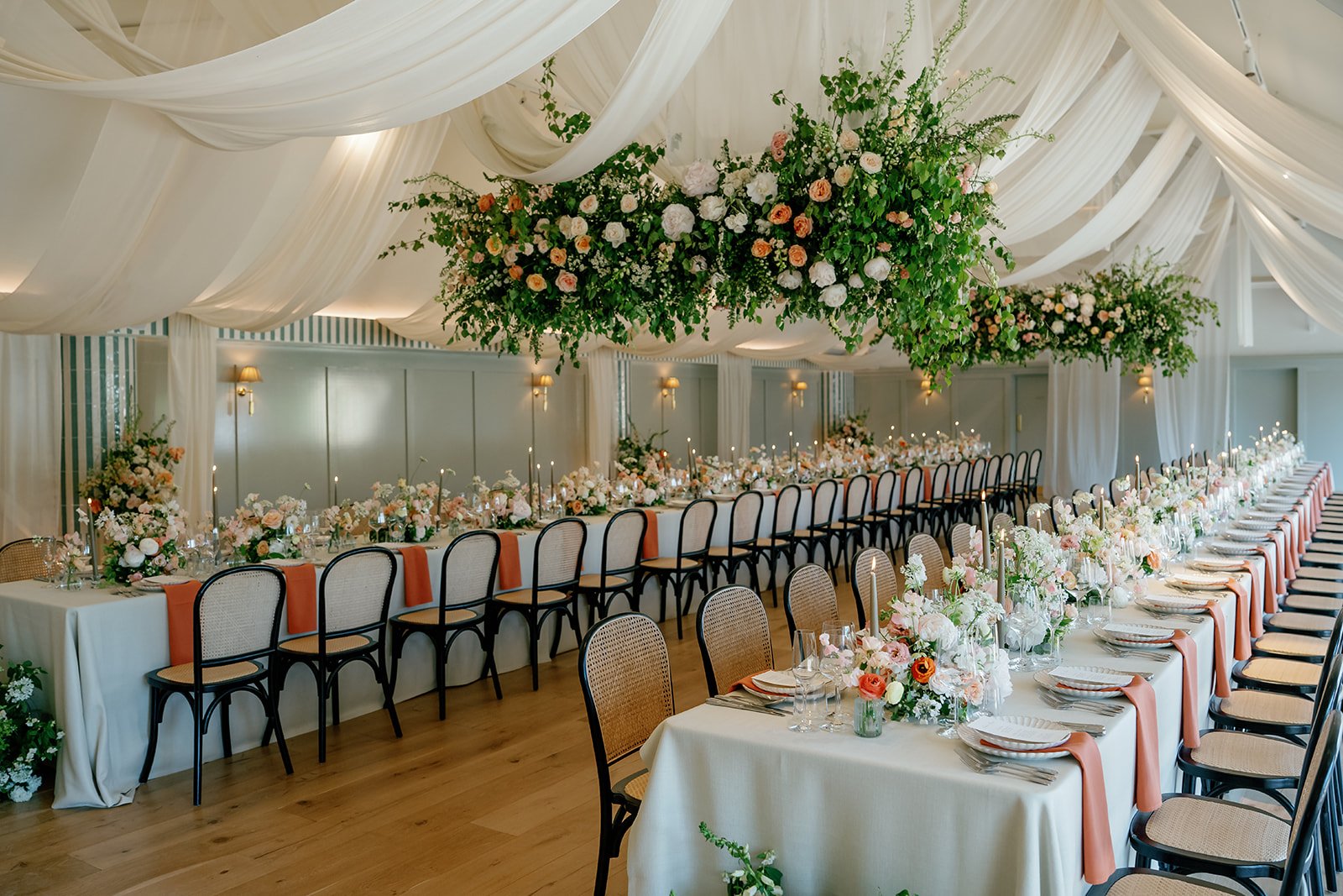 Elegant banquet hall decorated for a wedding or special event with long tables covered in white linen, adorned with pink, peach, and white floral arrangements, surrounded by chairs with beige cushions and peach sashes, and a large suspended floral ar