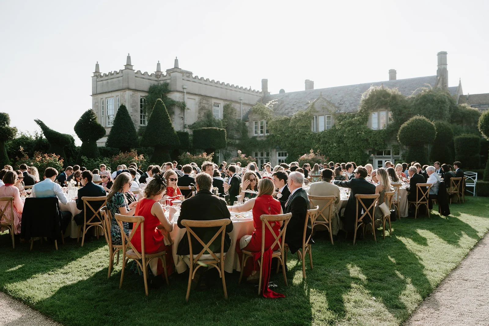 Guests dressed in formal attire seated at round tables on a well-manicured lawn during an outdoor event at a large historic estate with green hedges and a stone building in the background.