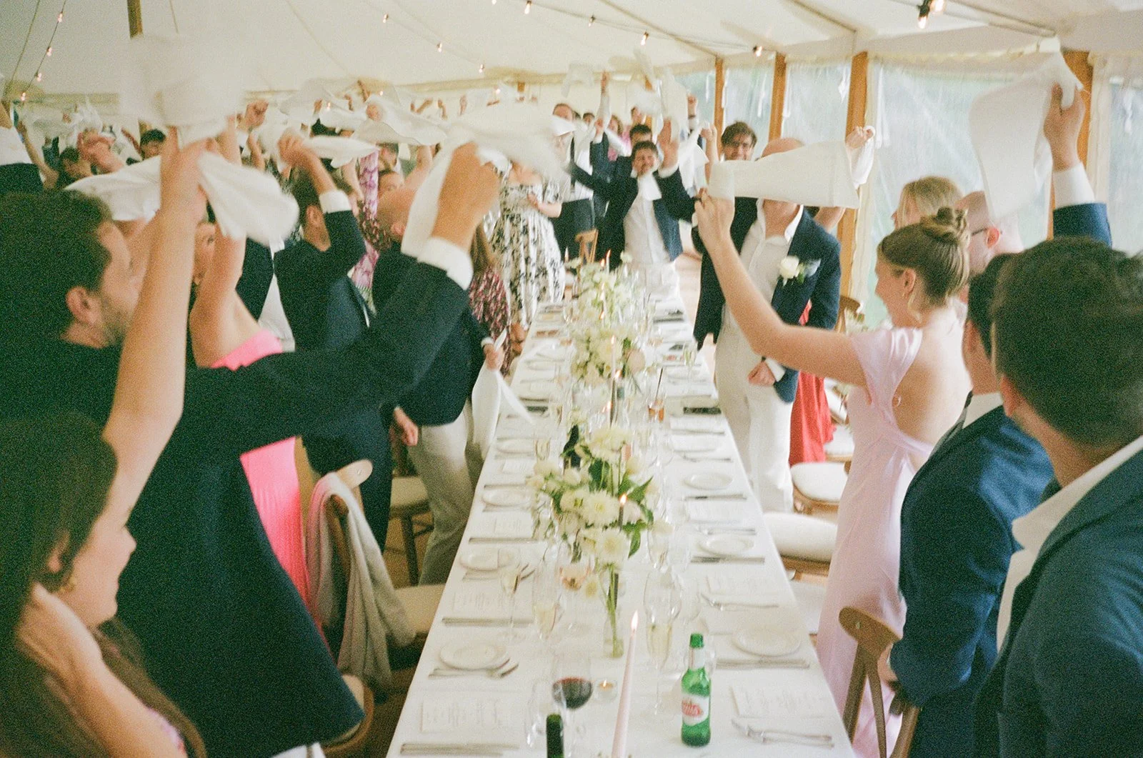 Guests standing and raising napkins during a wedding reception inside a decorated tent with a long table set with white flowers, candles, and place settings.