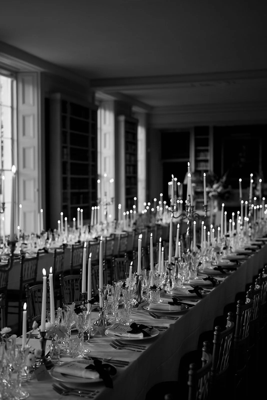 A long banquet table set with white tablecloth, candles, glassware, plates, silverware, and black ribbons in a dimly lit room with tall windows and bookshelves.