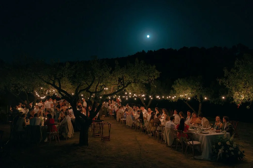 Outdoor nighttime wedding reception with long table, string lights, and guests under trees, illuminated by the moon.