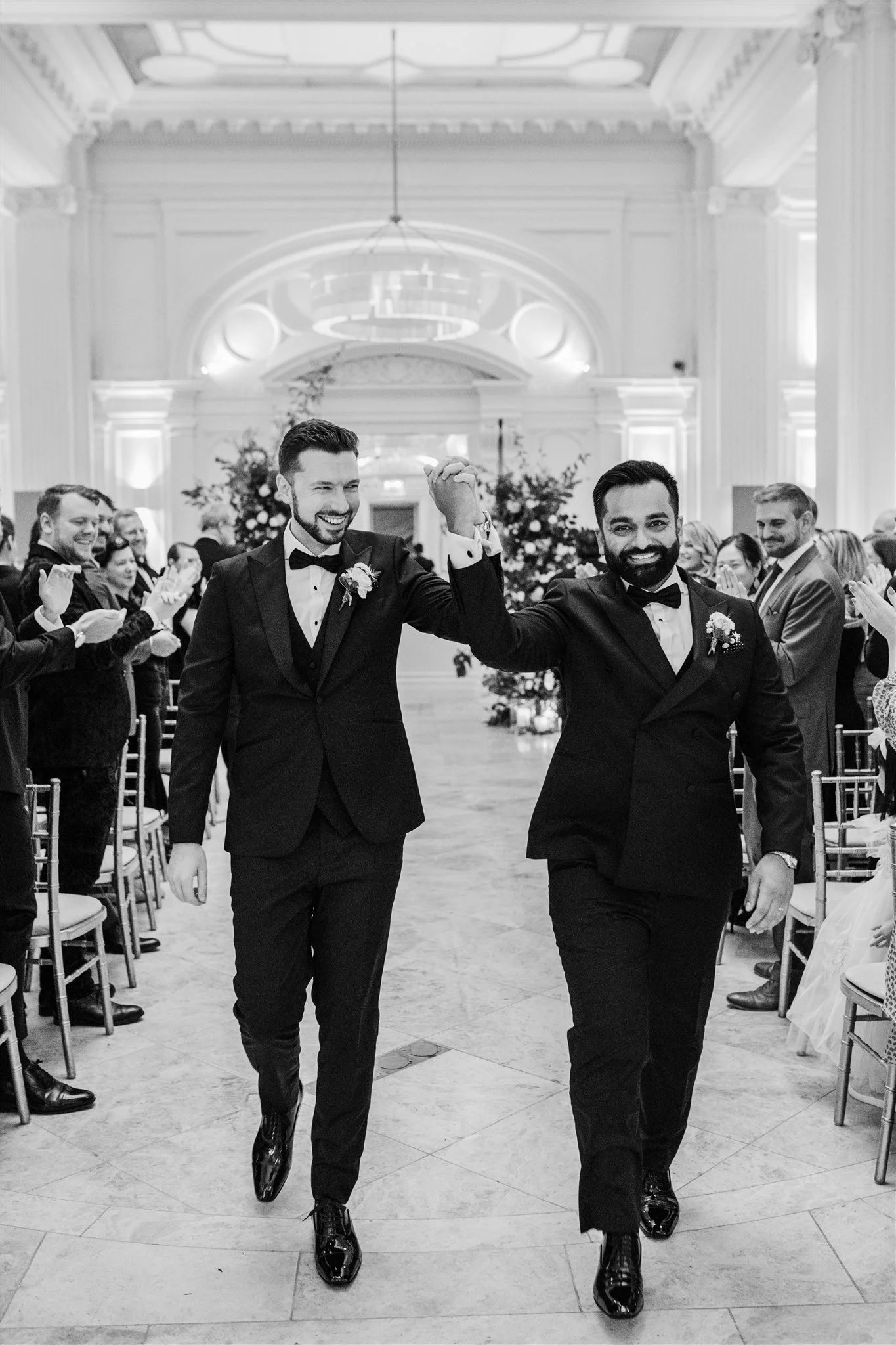 Two men in tuxedos walking down the aisle holding hands and smiling at the wedding ceremony, surrounded by seated guests clapping in a grand hall.